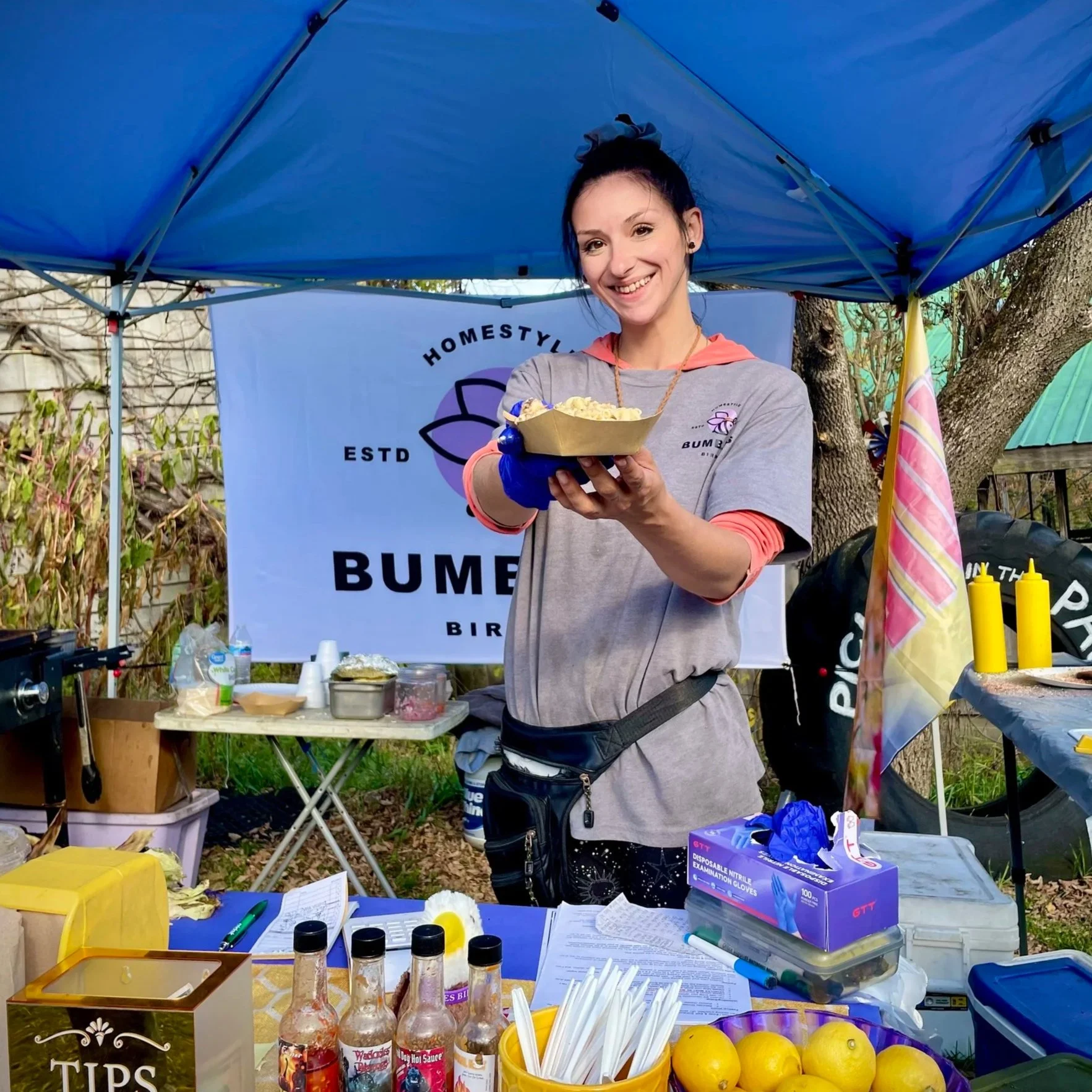 A smiling woman holding a serving of food at an outdoor food stand with a ‘Bumbles Birria' banner in the background. The stand has various condiments, lemons, and supplies on the table.