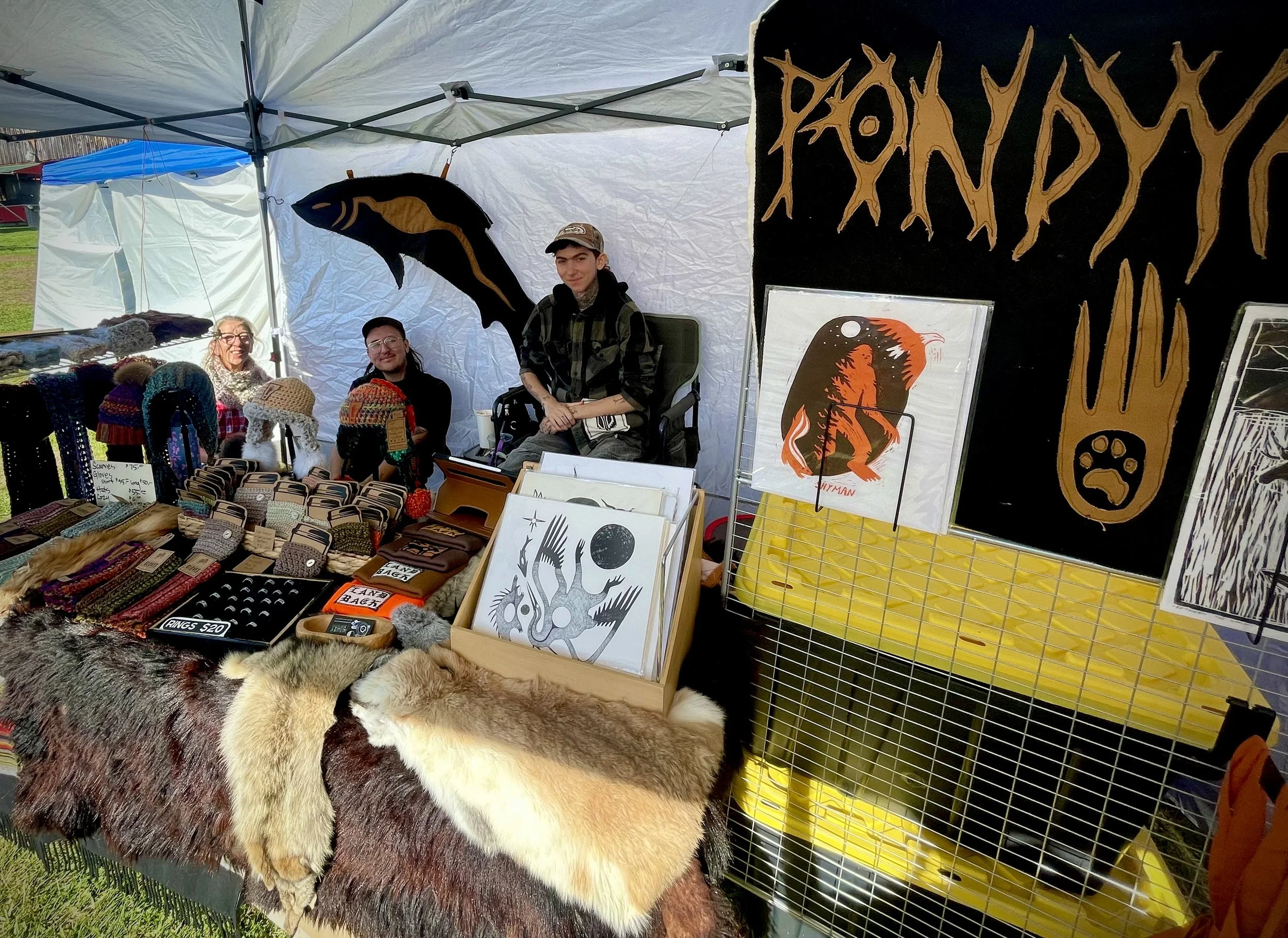 Market stall at an outdoor event selling handcrafted items, artwork, knitted hats, and animal pelts with three people sitting behind the display.