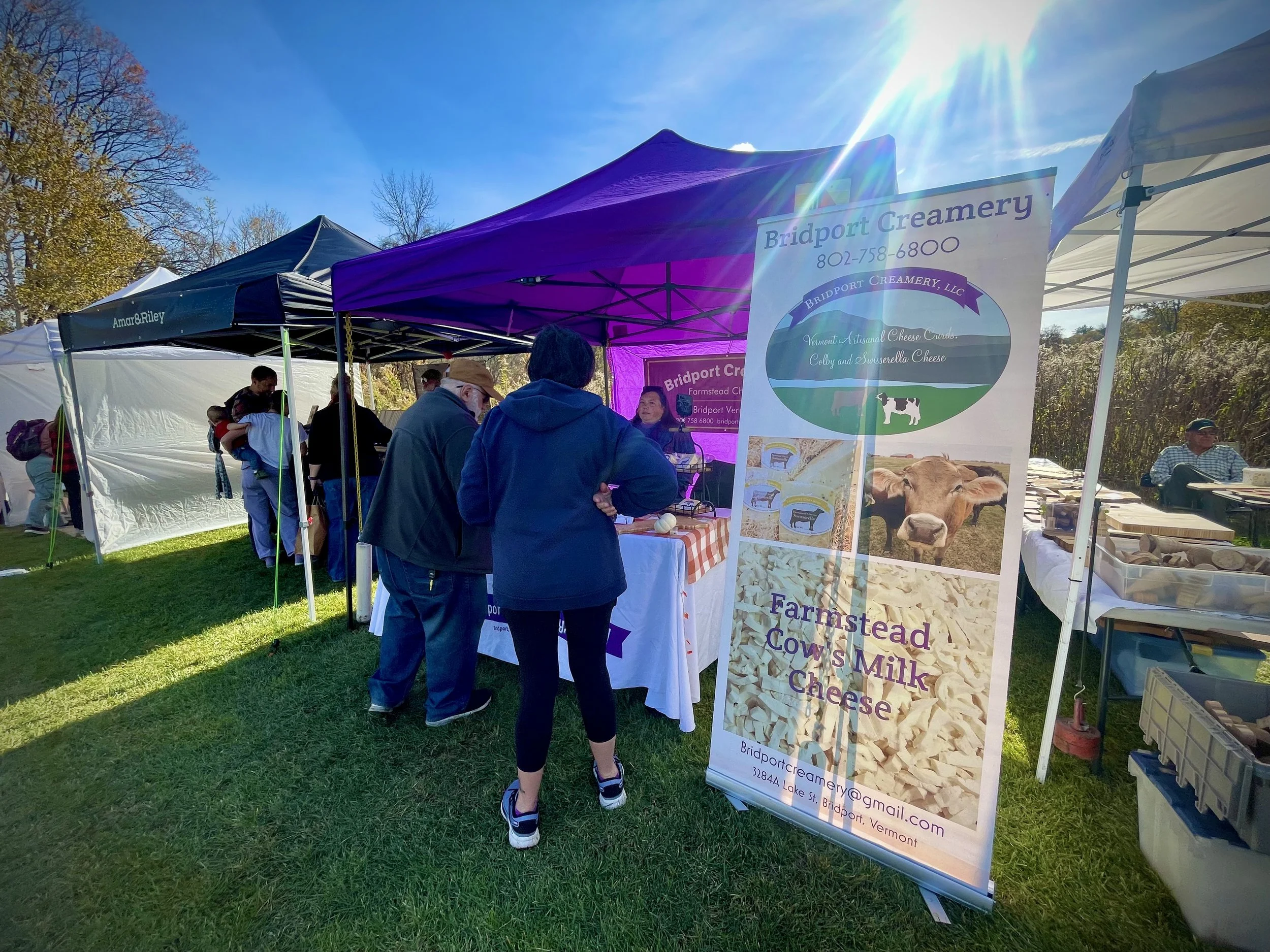 People shopping at a farm market booth with a large sign advertising Bridport Creamery's farmstead cow's milk cheese, with a cow image and contact information, under tents on a sunny day.