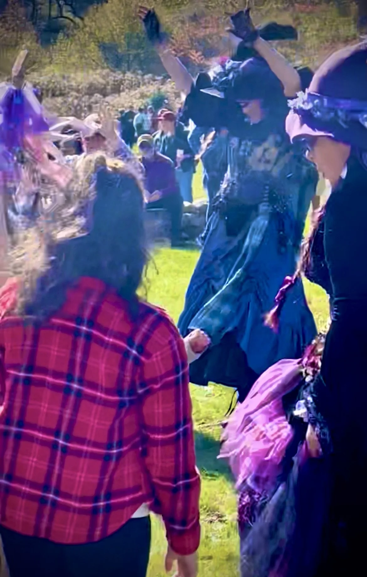 People in witch costumes dancing and holding hands in a circle outdoors during a daytime festival.