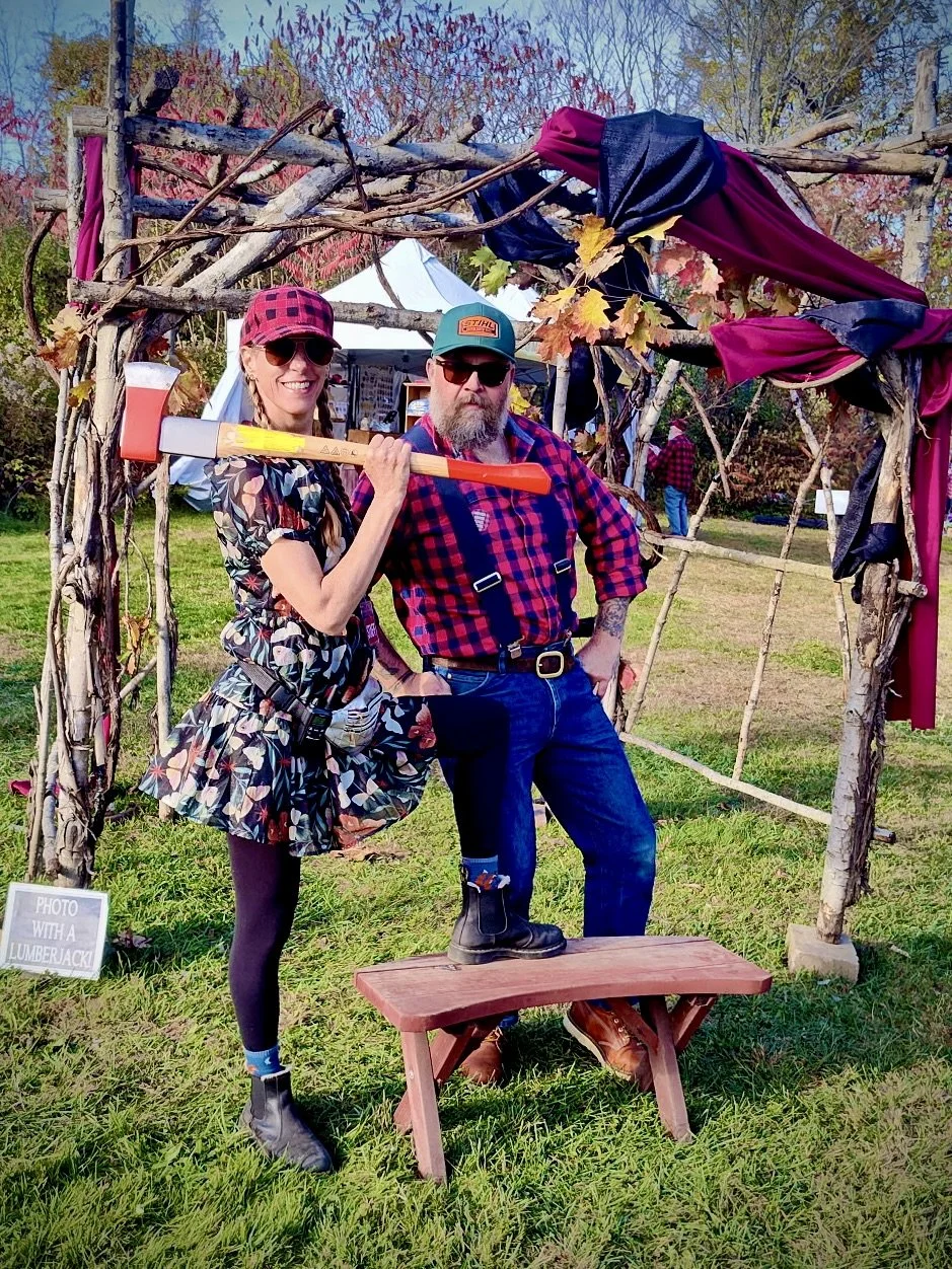 Two people standing under a rustic wooden arch decorated with fabric and leaves during an outdoor event. The woman on the left is smiling, wearing sunglasses, a floral dress, and a red checkered hat, holding a toy axe over her shoulder. The man on th