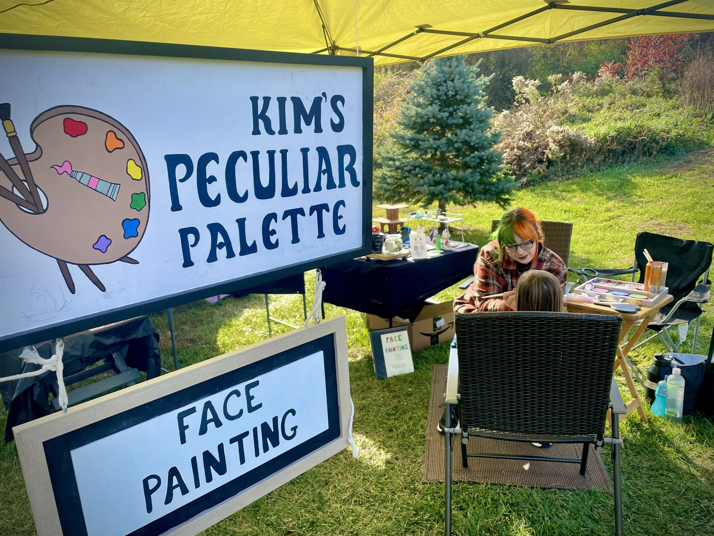 Artist creating face painting at an outdoor booth with signs reading 'Kim's Peculiar Palette' and 'Face Painting', set up under a yellow canopy in a grassy area with trees.