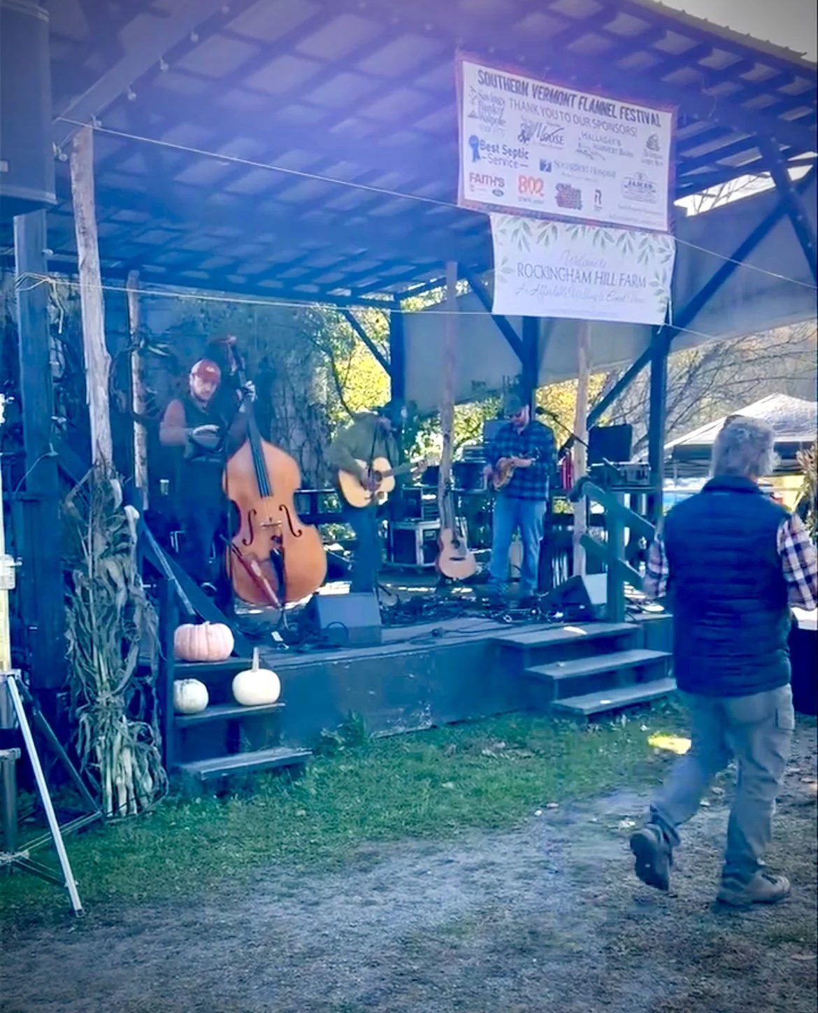 A small outdoor stage with three musicians performing; one playing a double bass, another with an acoustic guitar, and the third with a saxophone. There are pumpkins decorating the front of the stage. A man in a vest and work pants walks past, and th