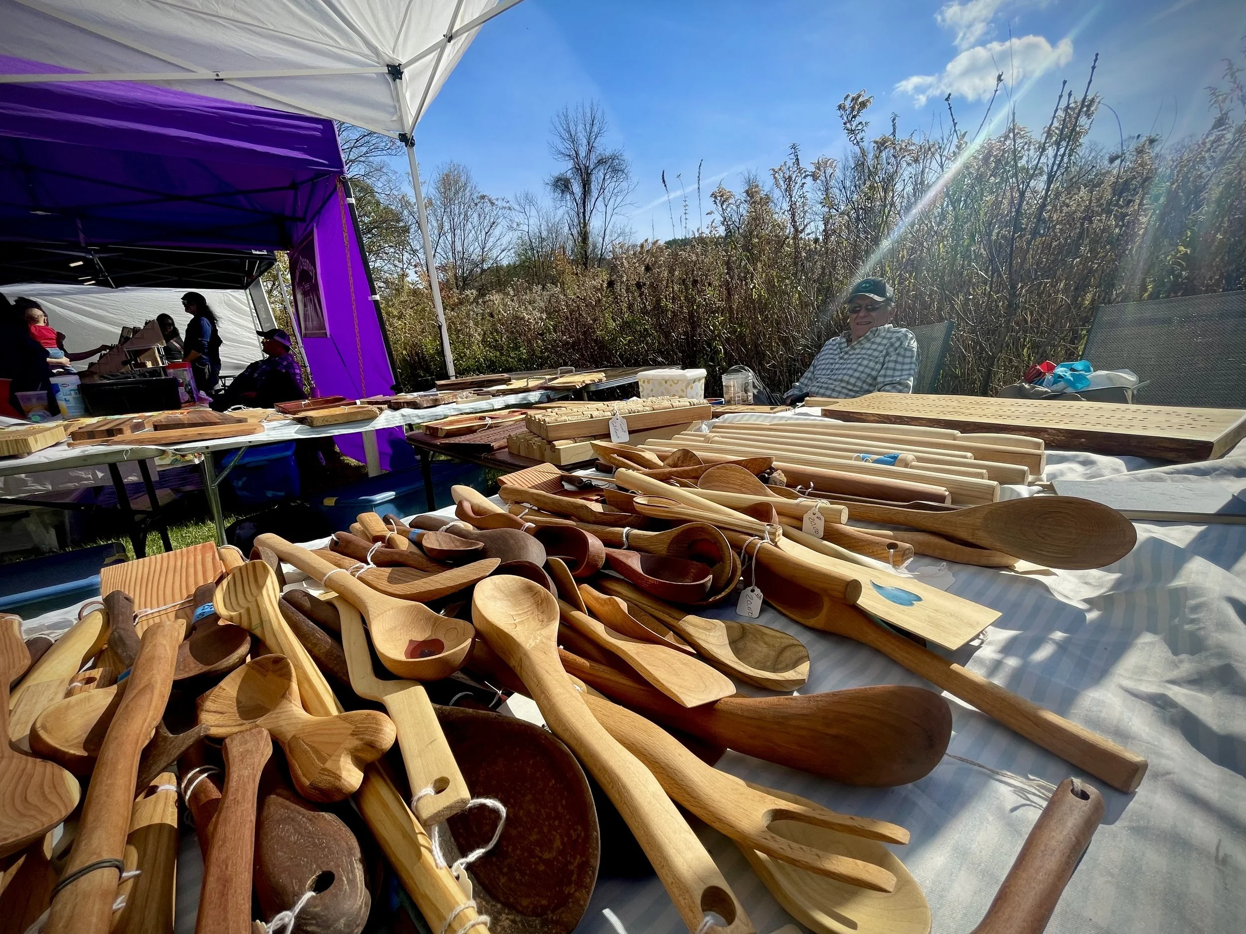 A craft fair outdoor booth displaying wooden spoons and kitchen utensils on a table. The background shows a person sitting in a chair and a group of people under a purple and white canopy with trees and blue sky above.