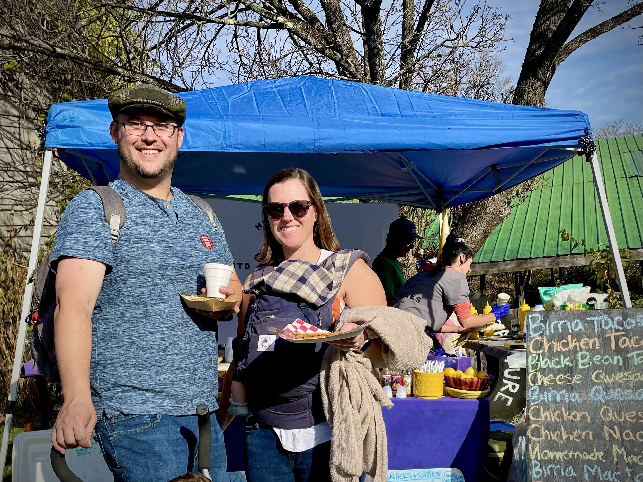 Two smiling people, a man and a woman, stand outdoors in front of a food stand with a blue canopy, at a fair or market on a sunny day. The man is holding a cup and plate, wearing glasses, a green cap, and a backpack. The woman is holding a plate and 