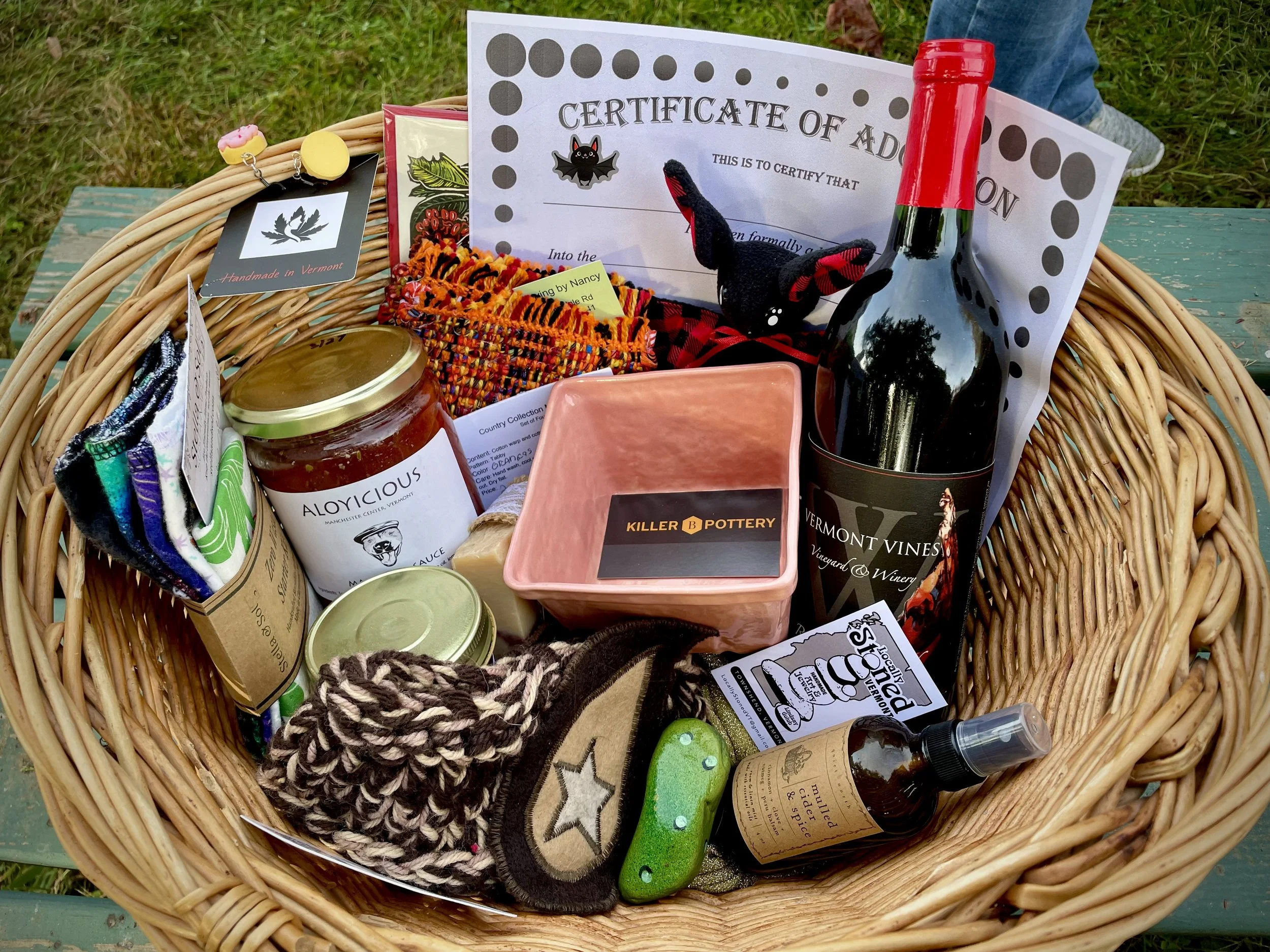A woven basket filled with various items including a jar of sauce labeled 'Aloicious', a pink ceramic dish, a bottle of Vermont Vines wine, a small plush toy, a jar of sauce, a spray bottle, colorful socks, a certificate, a small painted rock, and ot