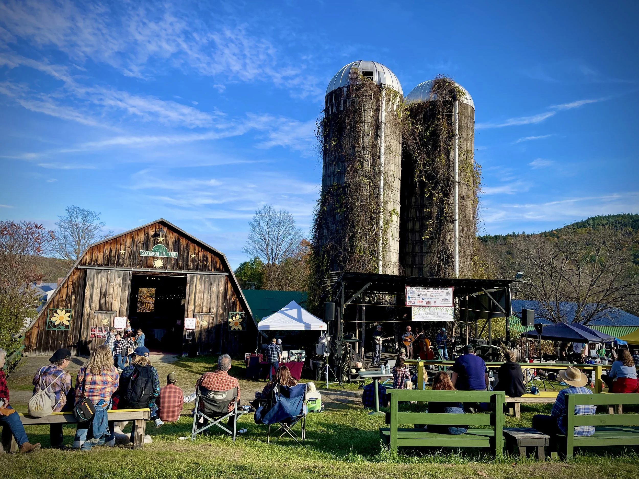 An outdoor concert at Rockingham Hill Farm with a rustic barn, a stage with musicians, and an audience sitting on benches and chairs on a grassy field under a clear blue sky.