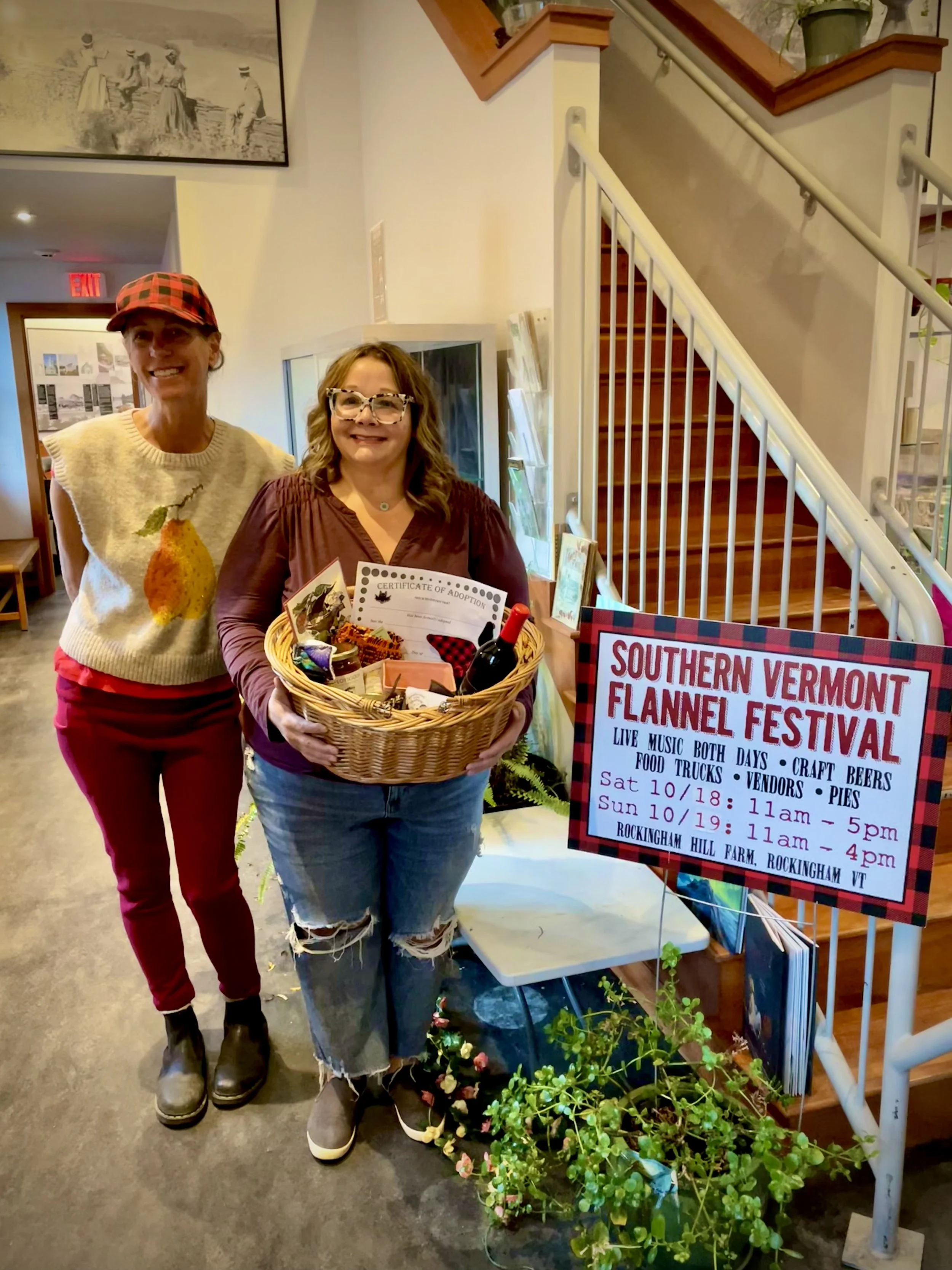 Two women standing inside a building near a staircase, one holding a gift basket with a certificate of adoption and a bottle of wine. A sign next to them promotes the Southern Vermont Flannel Festival, listing event details.