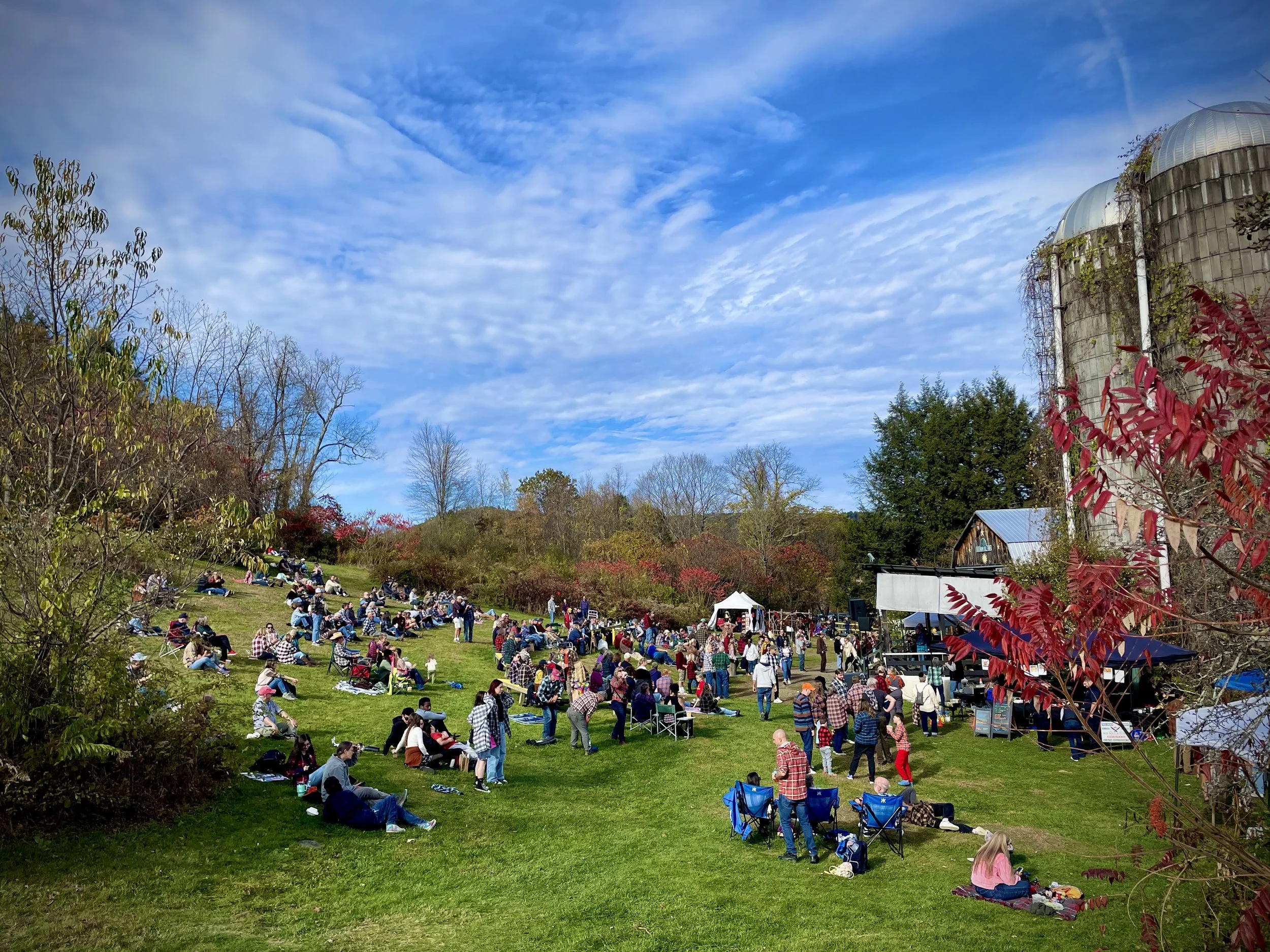Outdoor concert on a grassy hillside with many people sitting and standing, a stage with performers, trees with colorful fall foliage, and a blue sky with wispy clouds.