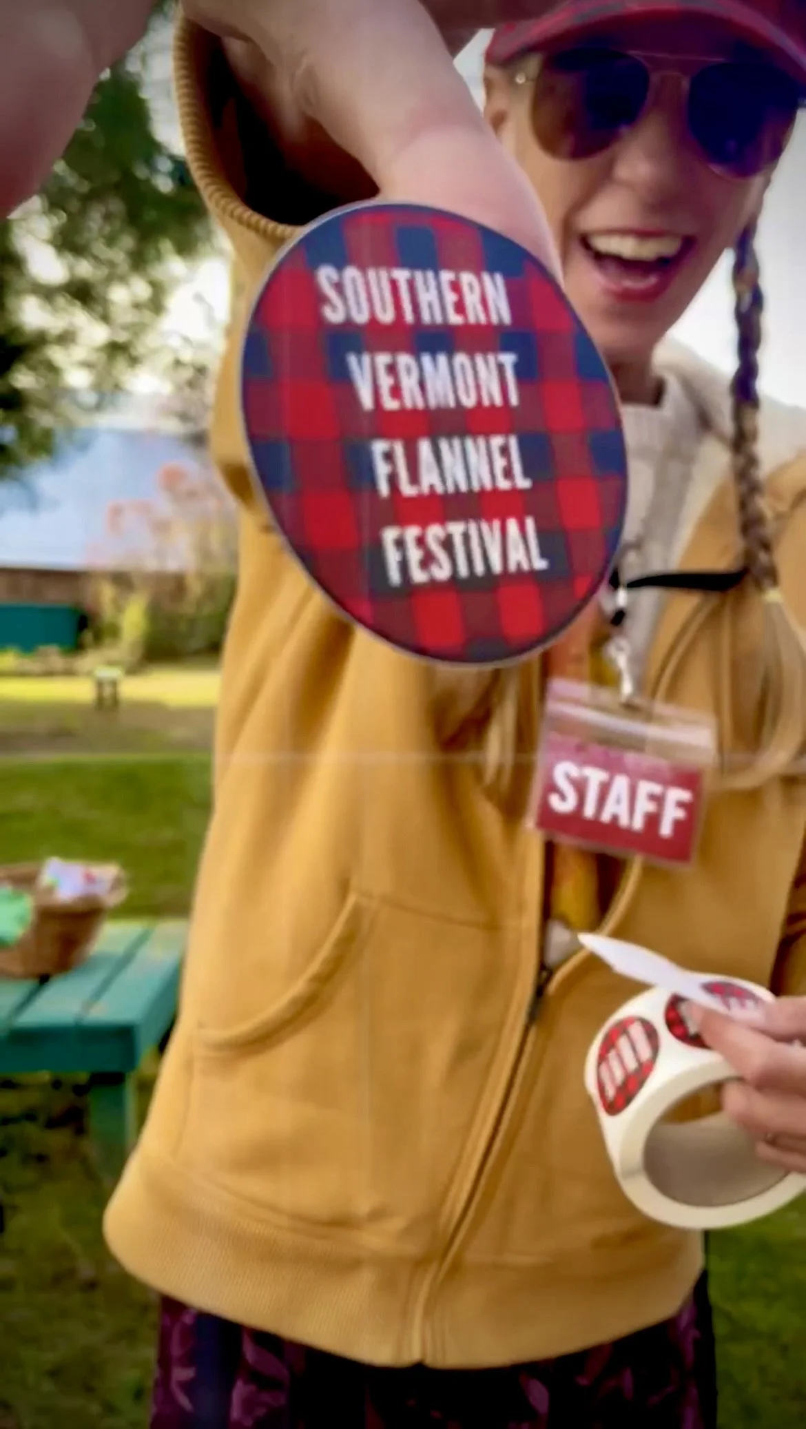 A smiling woman wearing sunglasses and a red buffalo plaid flannel cap, holding out a sticker with the text 'Southern Vermont Flannel Festival' and a roll of stickers, standing outdoors at a festival.