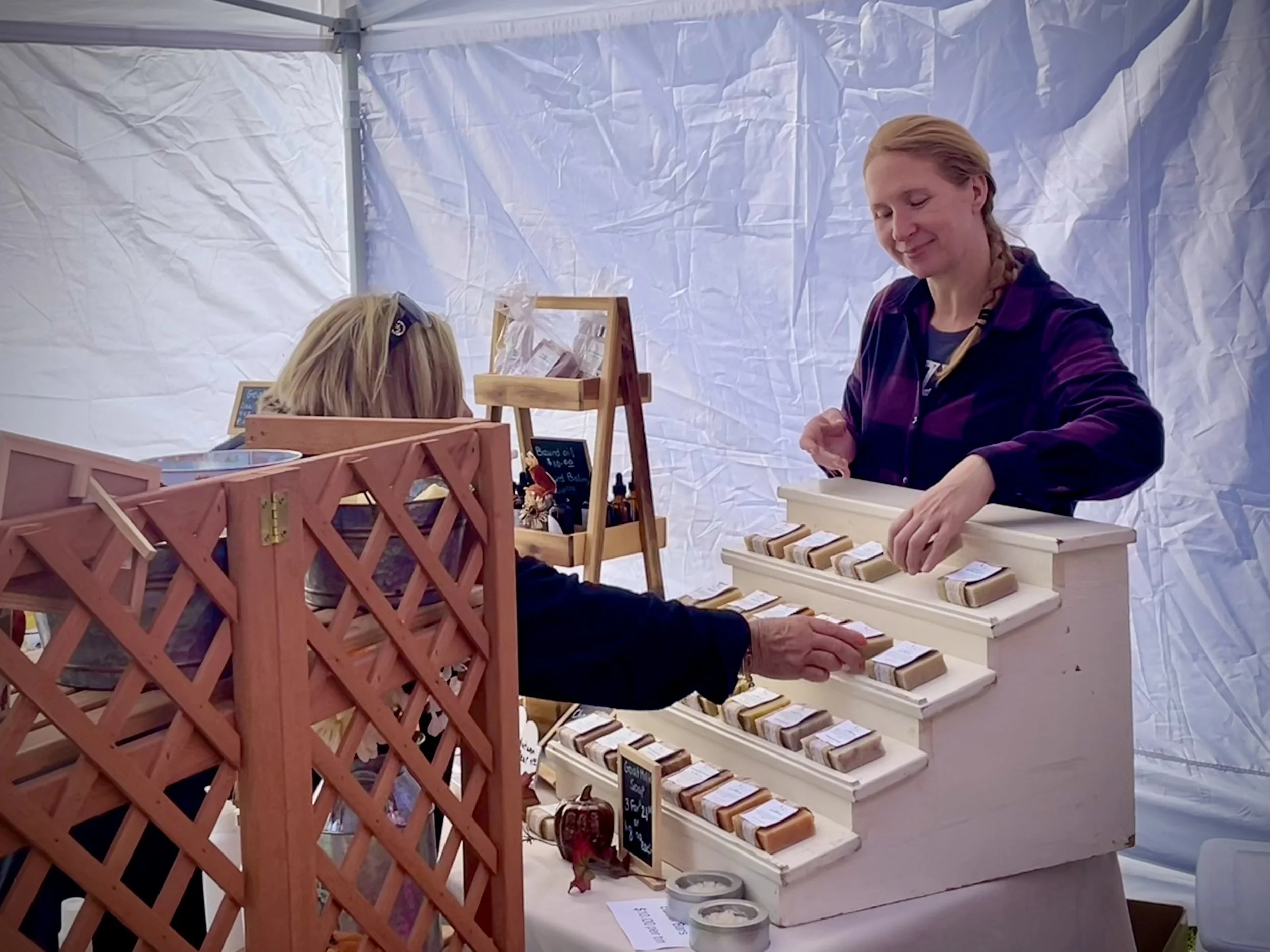 A woman selling soaps at a craft fair, handing a soap to a customer, with other soap products displayed on a white stand.