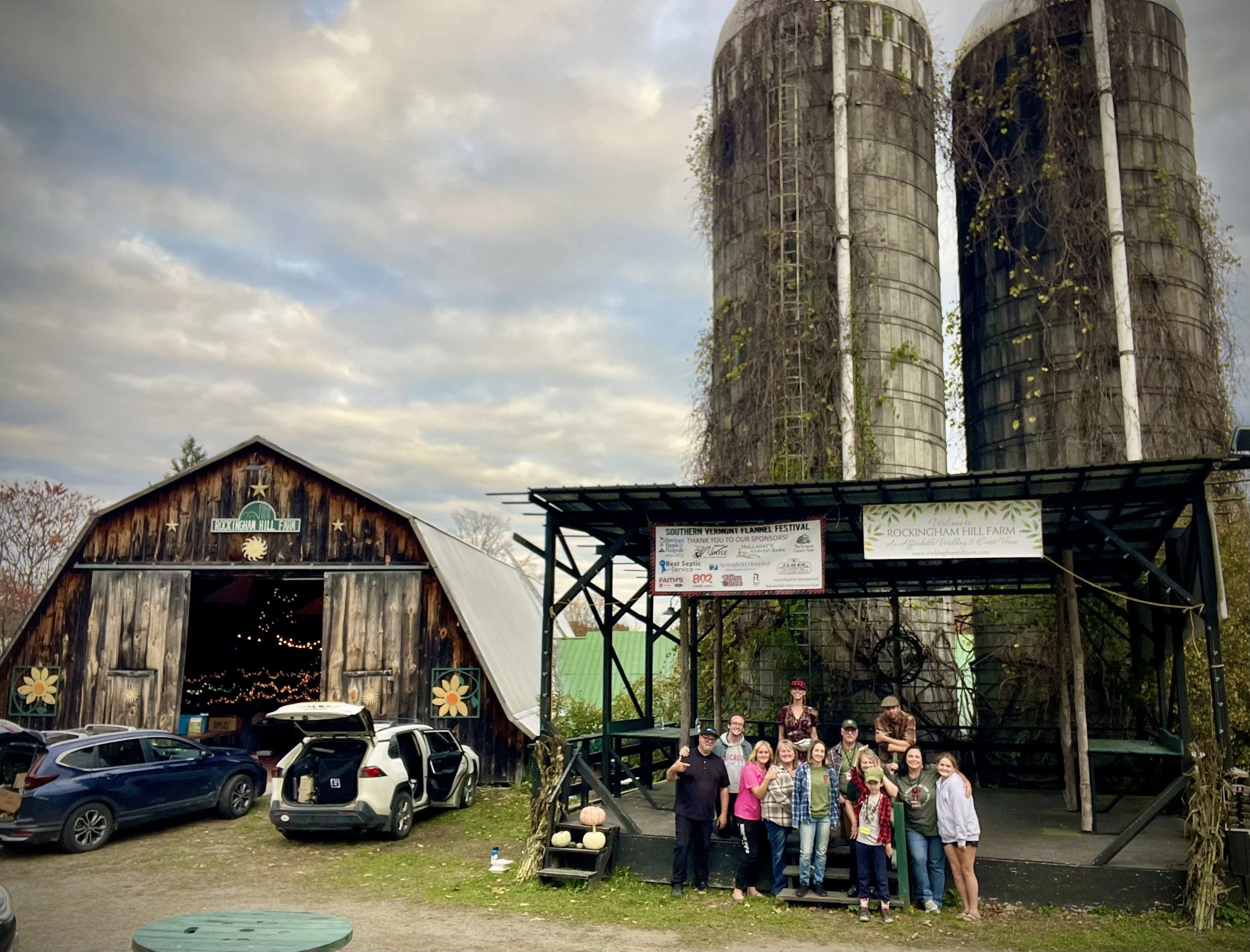 Volunteers pose on a stage in front of two large silo structures at Rockingham Hill Farm after the festival. A barn with open doors and string lights inside is visible on the left. Banners are displayed on the stage, and pumpkins are placed outside.