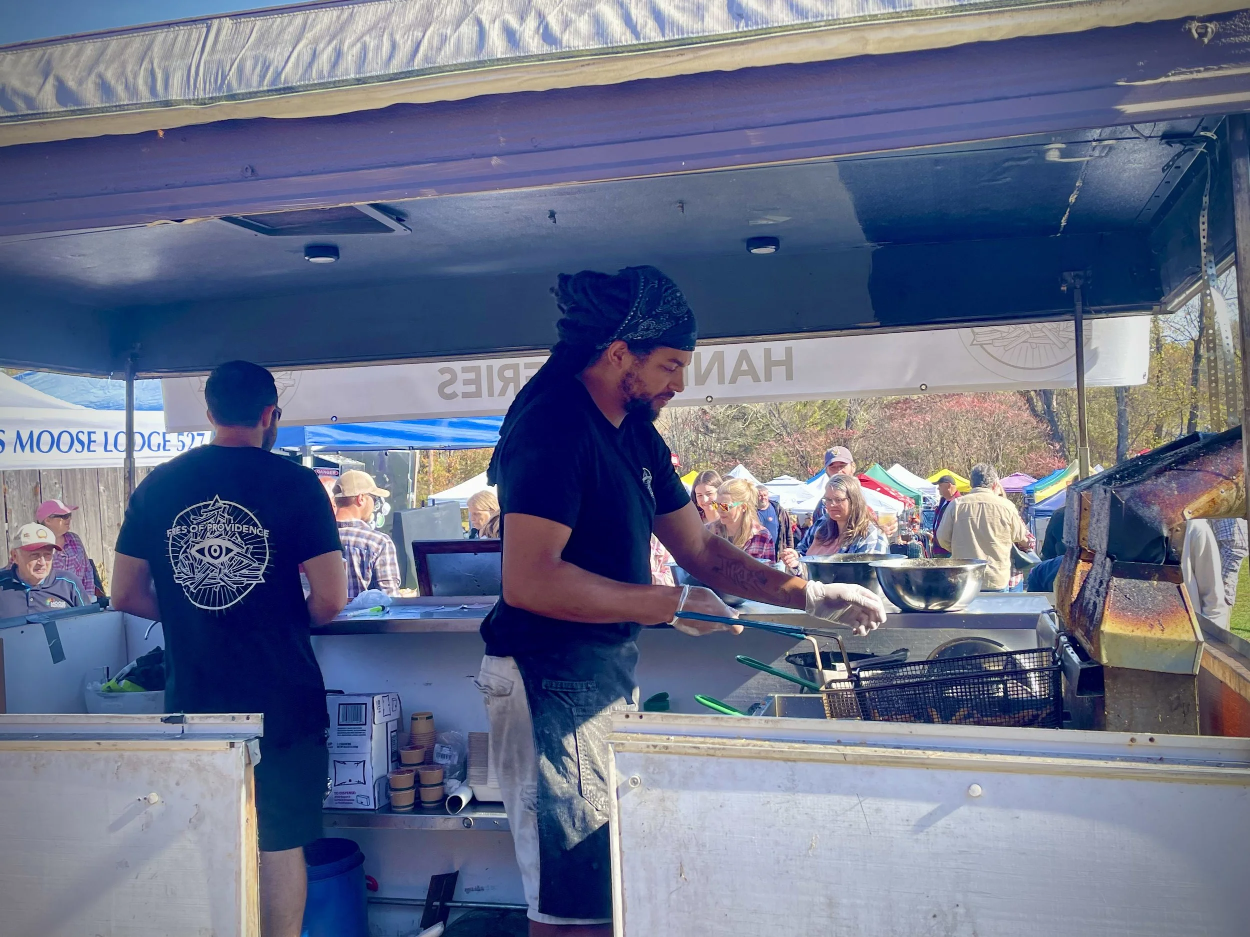 A man cooking food at an outdoor food stall during a festival, with people walking and shopping in the background.