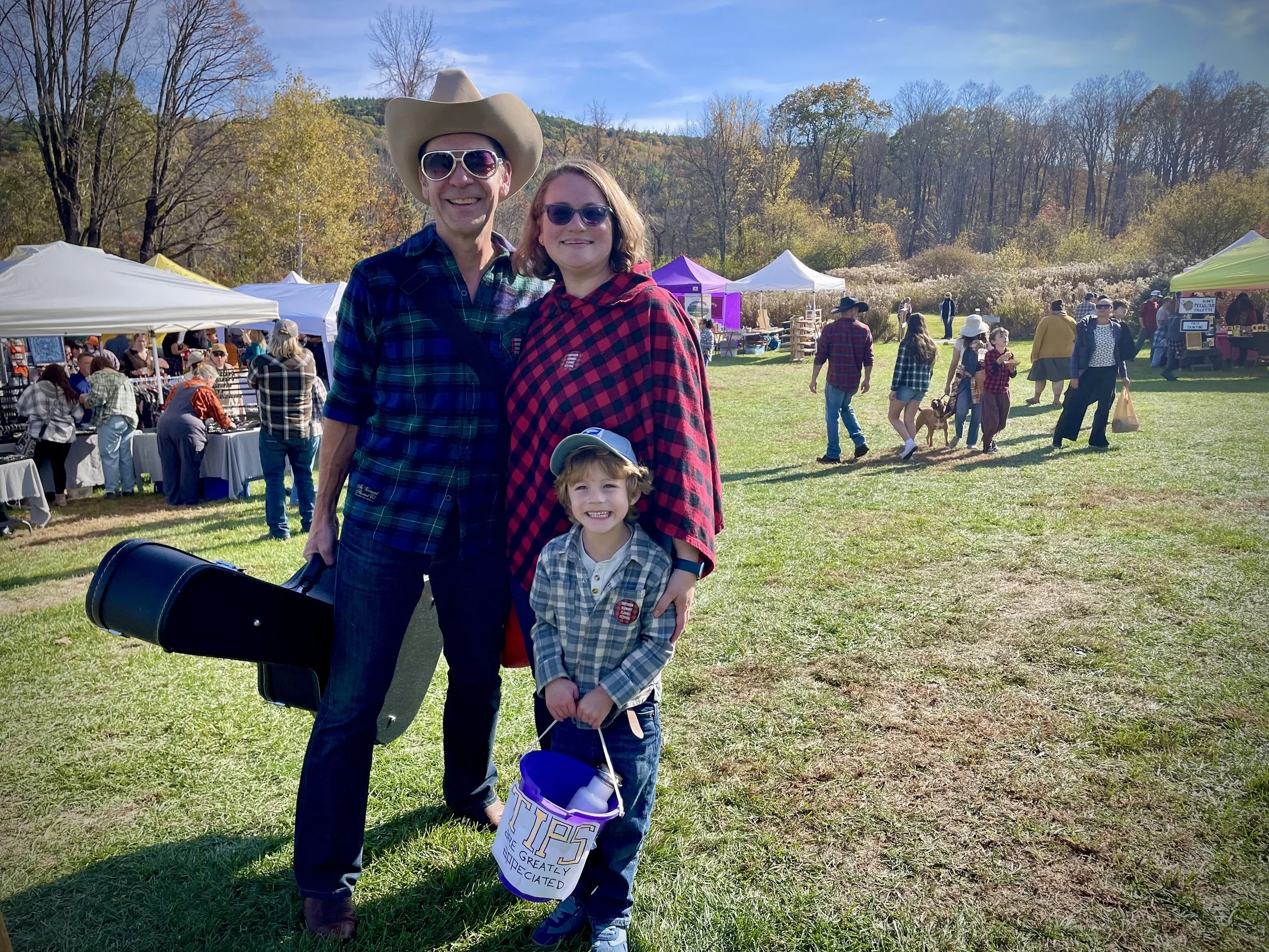 Family of 3 outdoors at a festival, tents and people in the background. The man wears a cowboy hat, Elvis sunglasses, and plaid shirt; the woman wears sunglasses and a plaid shawl; the boy wears a cap, plaid shirt, and is holding a bucket of candy.
