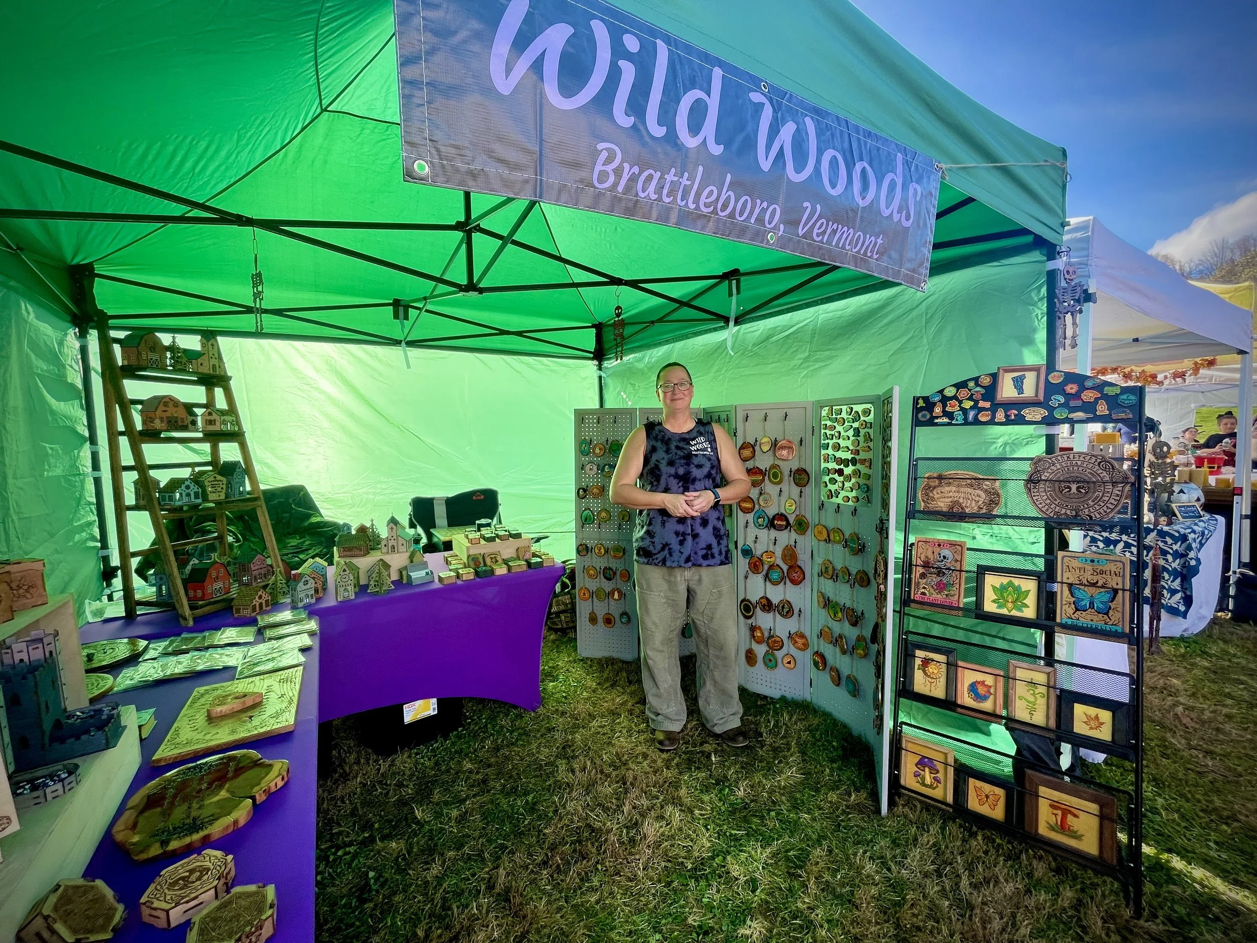A woman standing inside a green outdoor market booth with handmade crafts including wood carvings, painted tiles, and jewelry. The booth has a sign that reads 'Wild Woods, Brattleboro, Vermont' and is set up at an outdoor event with other booths visi