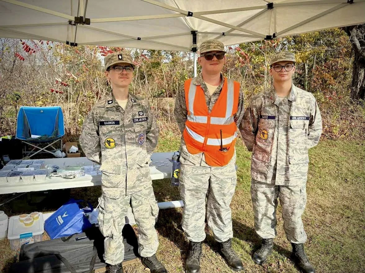 Three Civil Air Patrol Cadets standing under a tent, two women and one man, all in camouflage uniforms. The man in the middle is wearing an orange safety vest. There is a table with supplies behind them and trees with red leaves in the background.