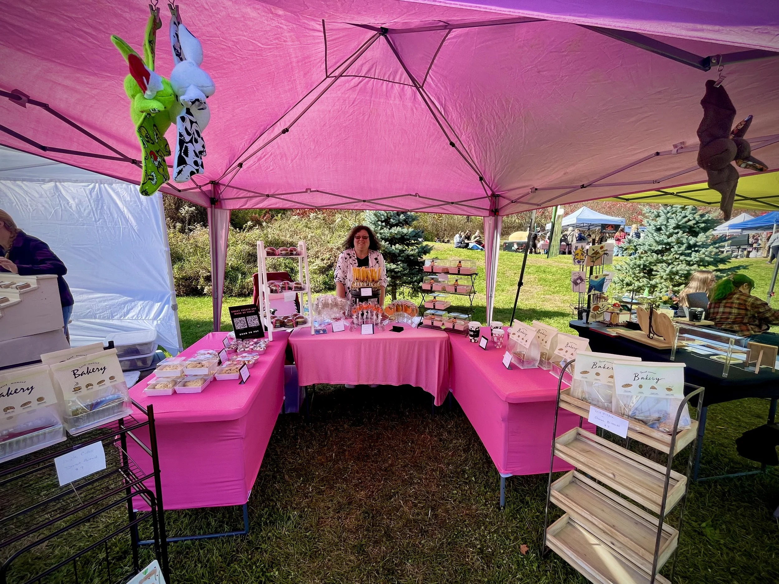 A woman stands behind a pink table at an outdoor market stall, selling handmade items under a pink canopy. Stuffed bunnies are hanging from the canopy, and there are other vendors and trees in the background.