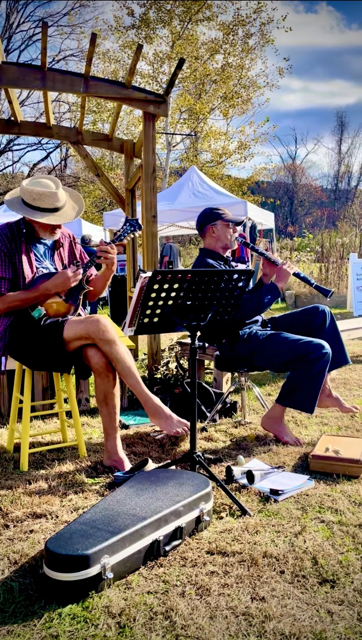 Two musicians perform outdoors on a sunny day, one playing a ukulele and the other playing a clarinet, with a tent and trees in the background. Both are barefoot, with music equipment around them.