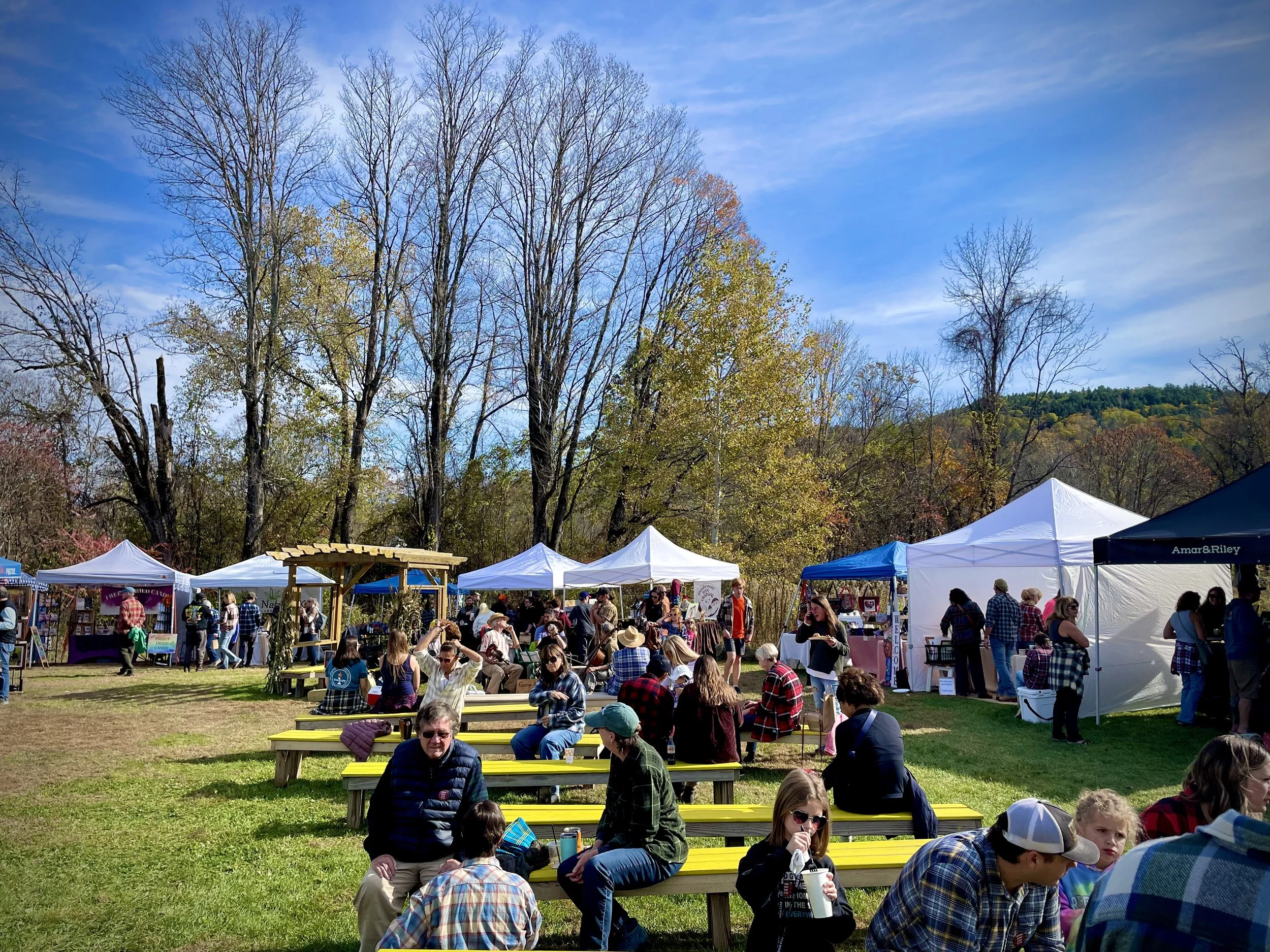People attending an outdoor market with tents and booths, sitting on yellow benches under a clear blue sky surrounded by trees.