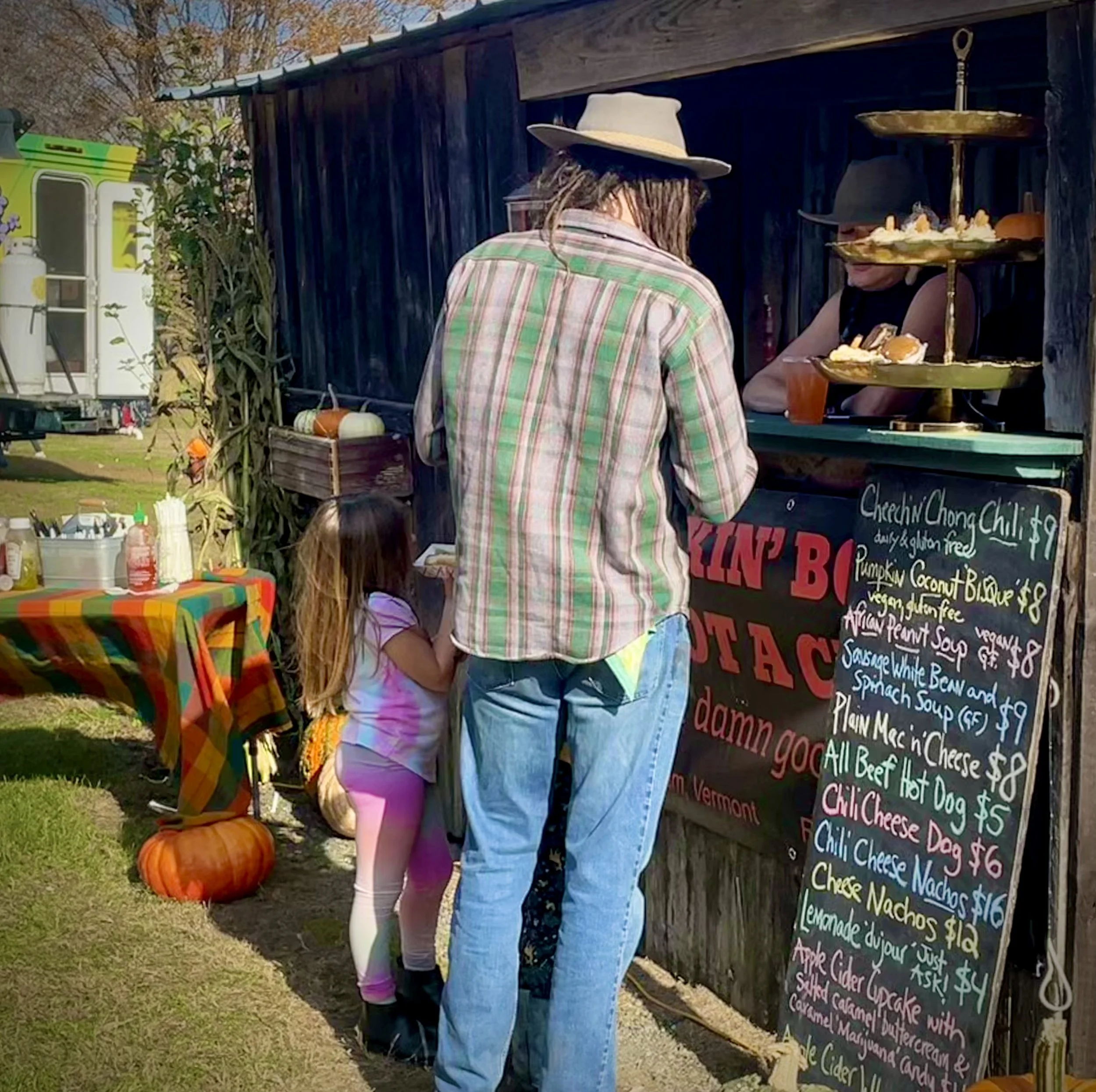 People standing at an outdoor food stand with a chalkboard menu, pumpkin decorations, and a table with condiments