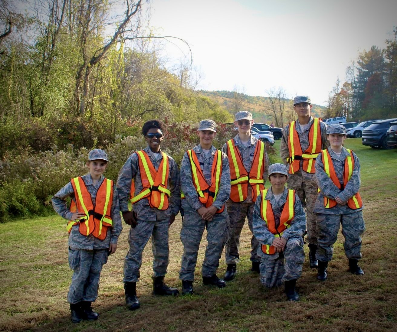 Group of seven young Civil Air Patrol Cadets in camouflage uniform and reflective safety vests posing outdoors while volunteering as festival parking attendants, with trees and parked cars in the background.