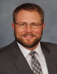 Portrait of a man with glasses, a beard, and short brown hair, wearing a dark suit, white shirt, and patterned tie, smiling against a blue background.