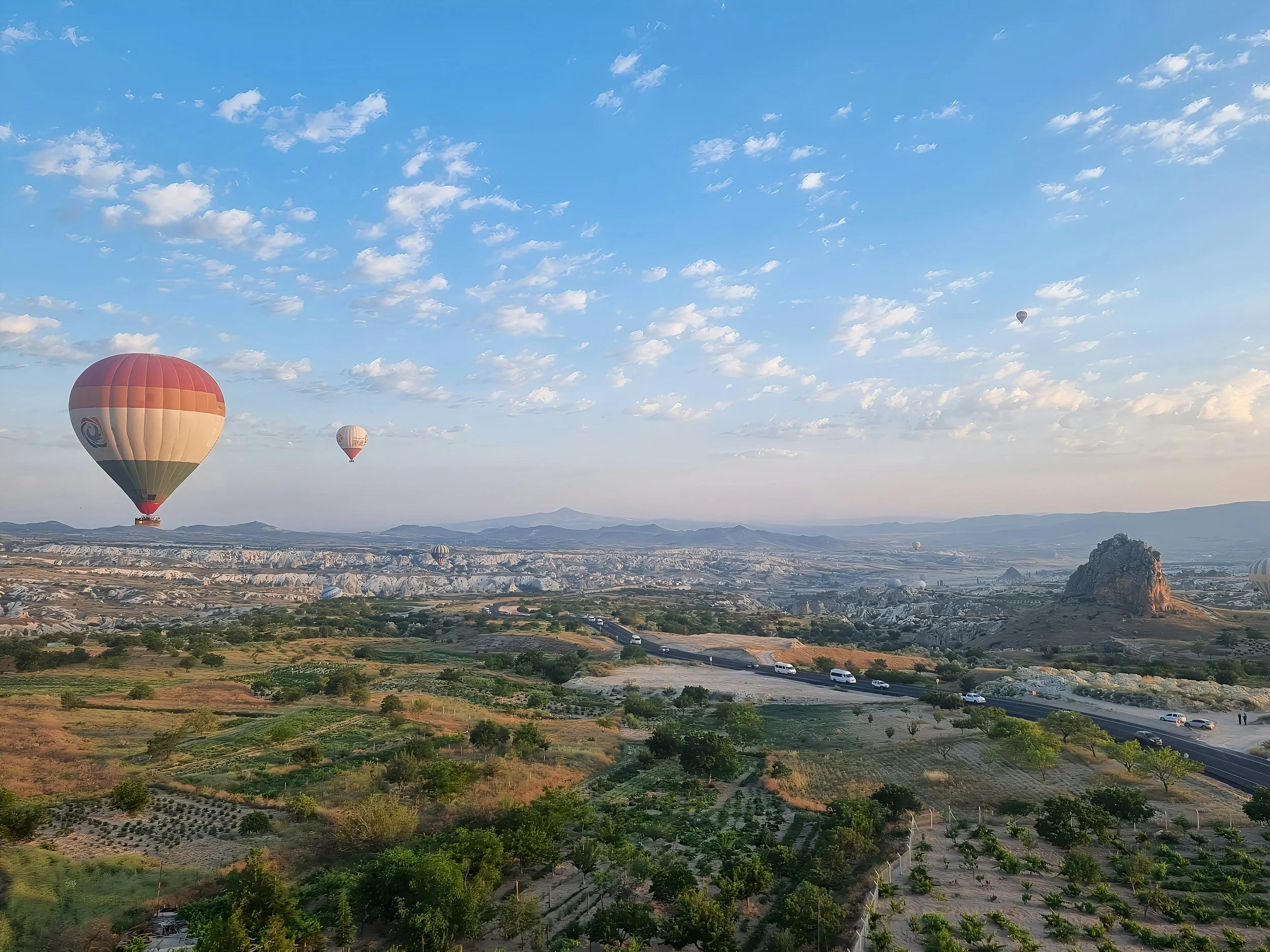 Landscape with multiple hot air balloons floating in a clear sky over a rural area with green fields and unique rock formations.