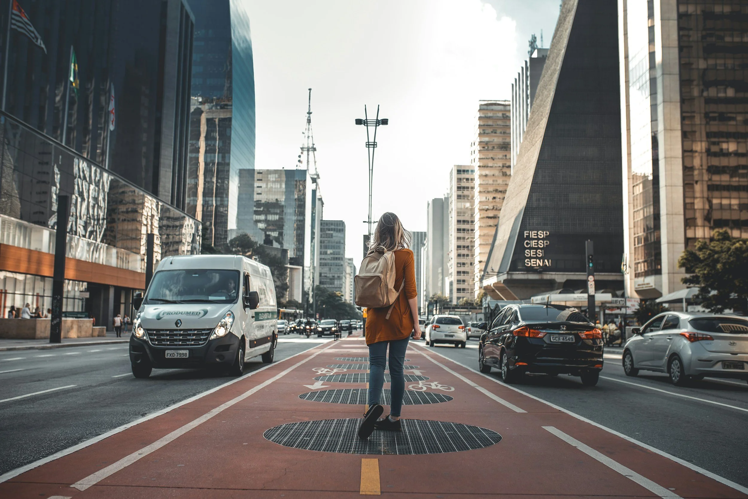 A woman with a backpack walking across a city bike lane on a busy street with tall buildings, cars, and a clear sky.