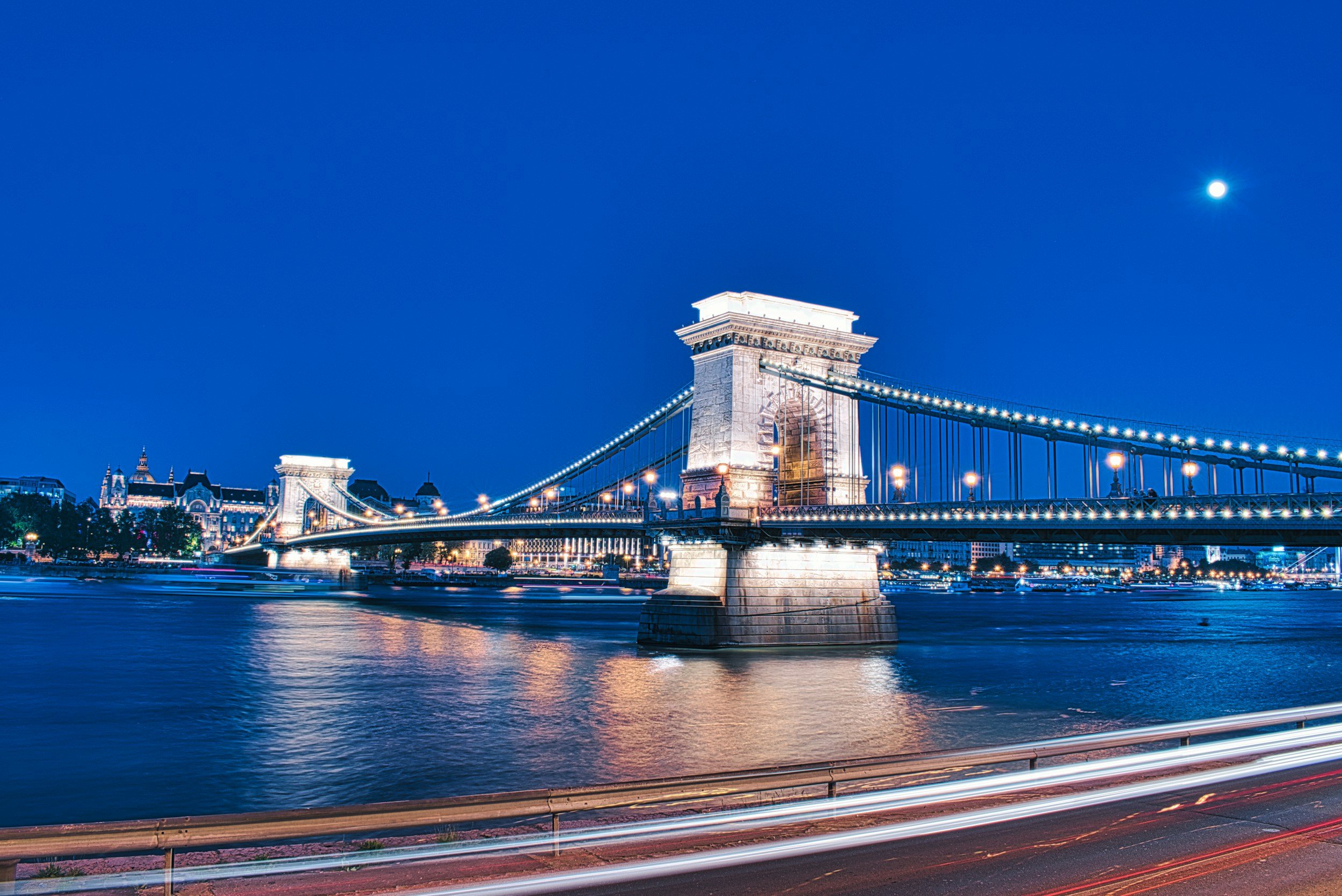Nighttime view of the Chain Bridge in Budapest, Hungary, illuminated with lights, across the Danube River under a clear sky with the moon overhead.