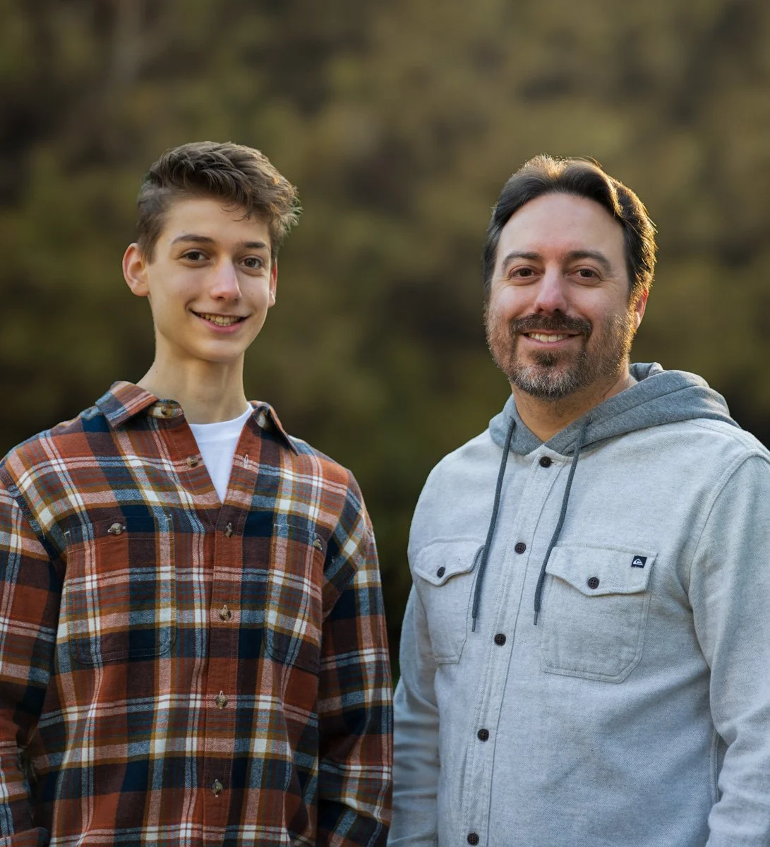 A young man and an older man smiling outdoors with blurred trees or forest in the background.