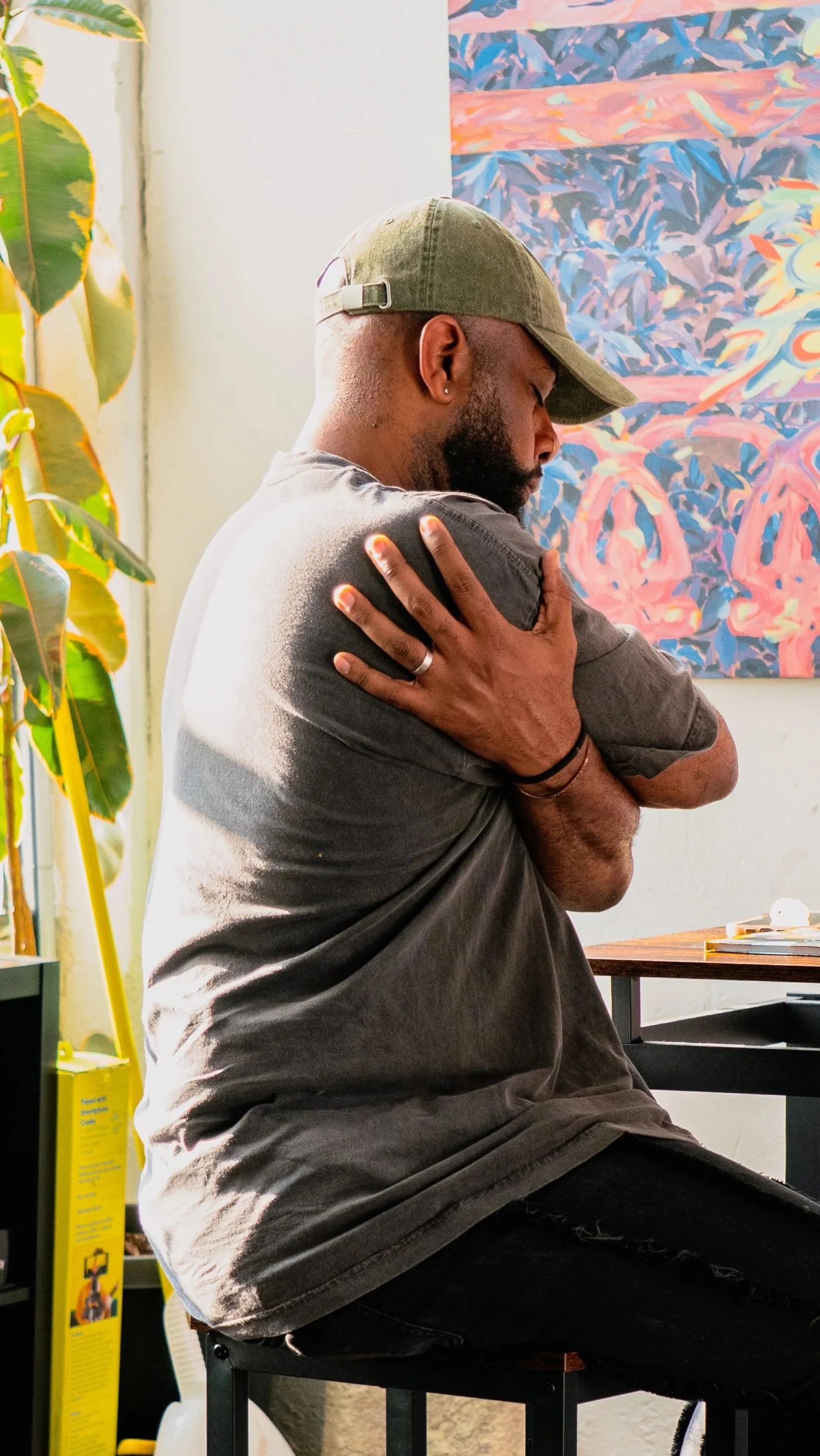 A man with a beard and a baseball cap sitting on a black stool in a room, hugging himself with his arms crossed over his chest. There is a yellow mop and other cleaning supplies next to a green plant, and colorful artwork hanging on the white wall be