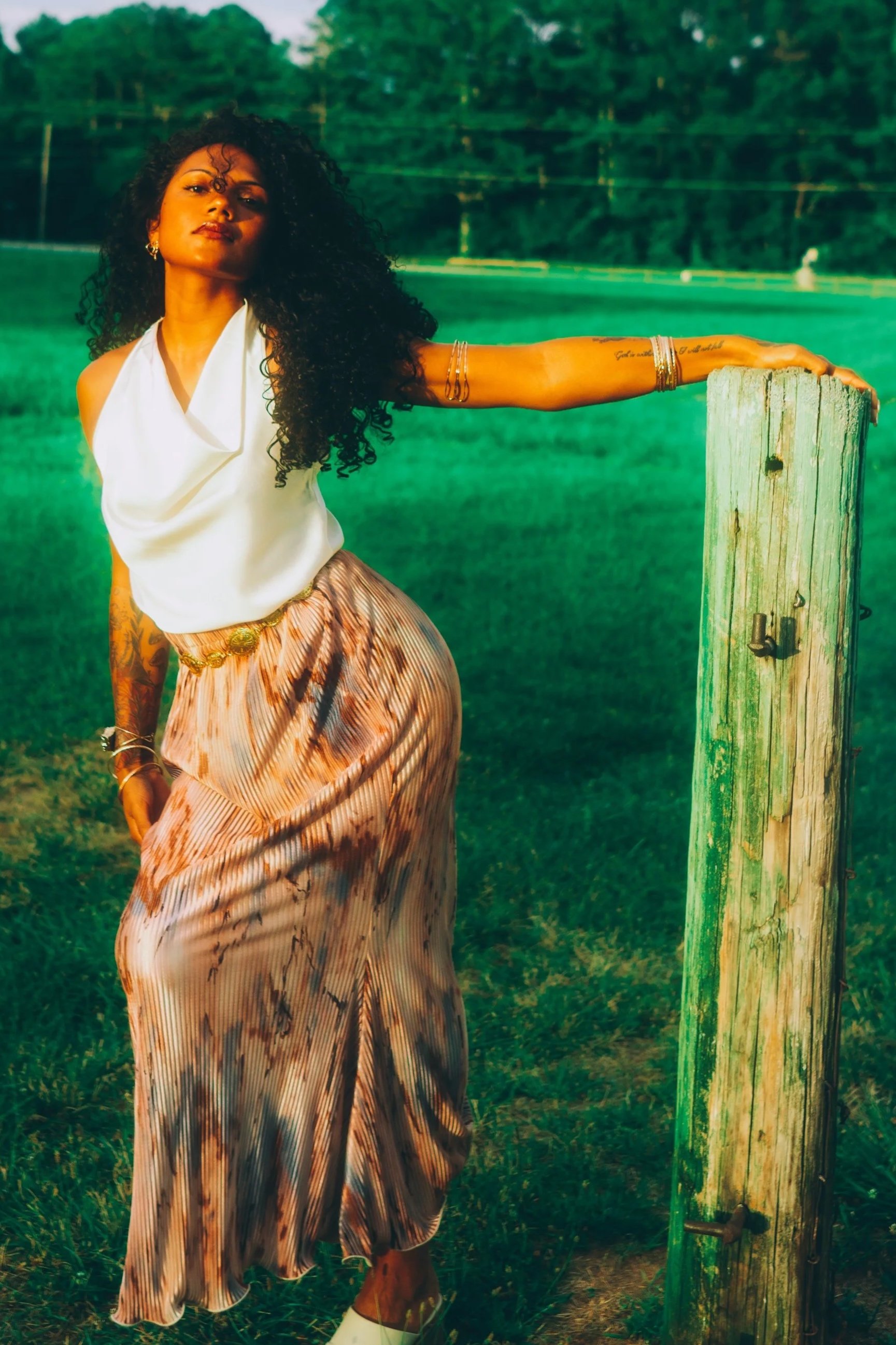 A woman with curly hair standing in a grassy field, leaning on a wooden fence post, wearing a white sleeveless top and a long, patterned skirt.