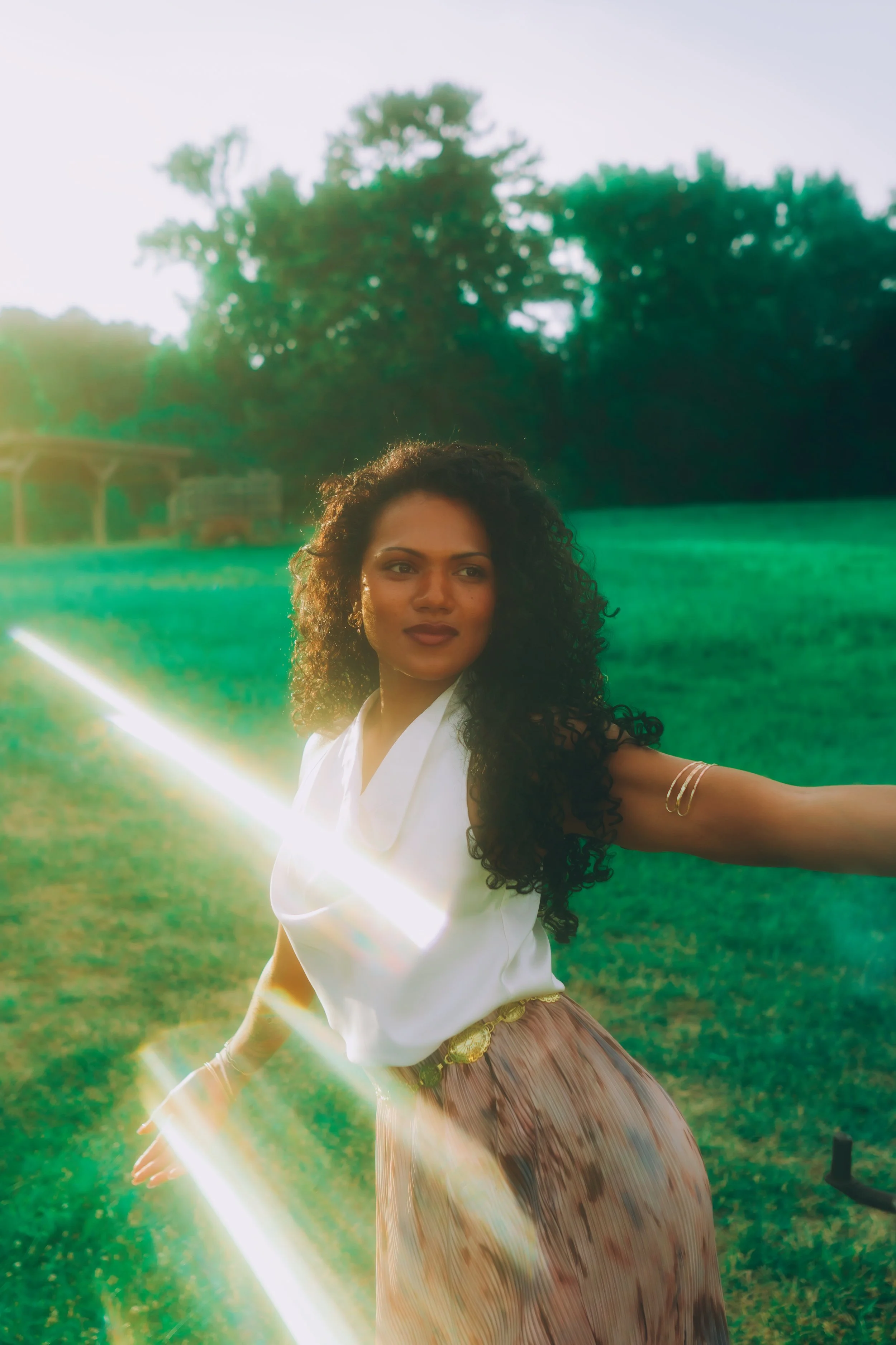A woman with curly hair standing outdoors in a green field during the daytime, with sunlight creating lens flare in the image.
