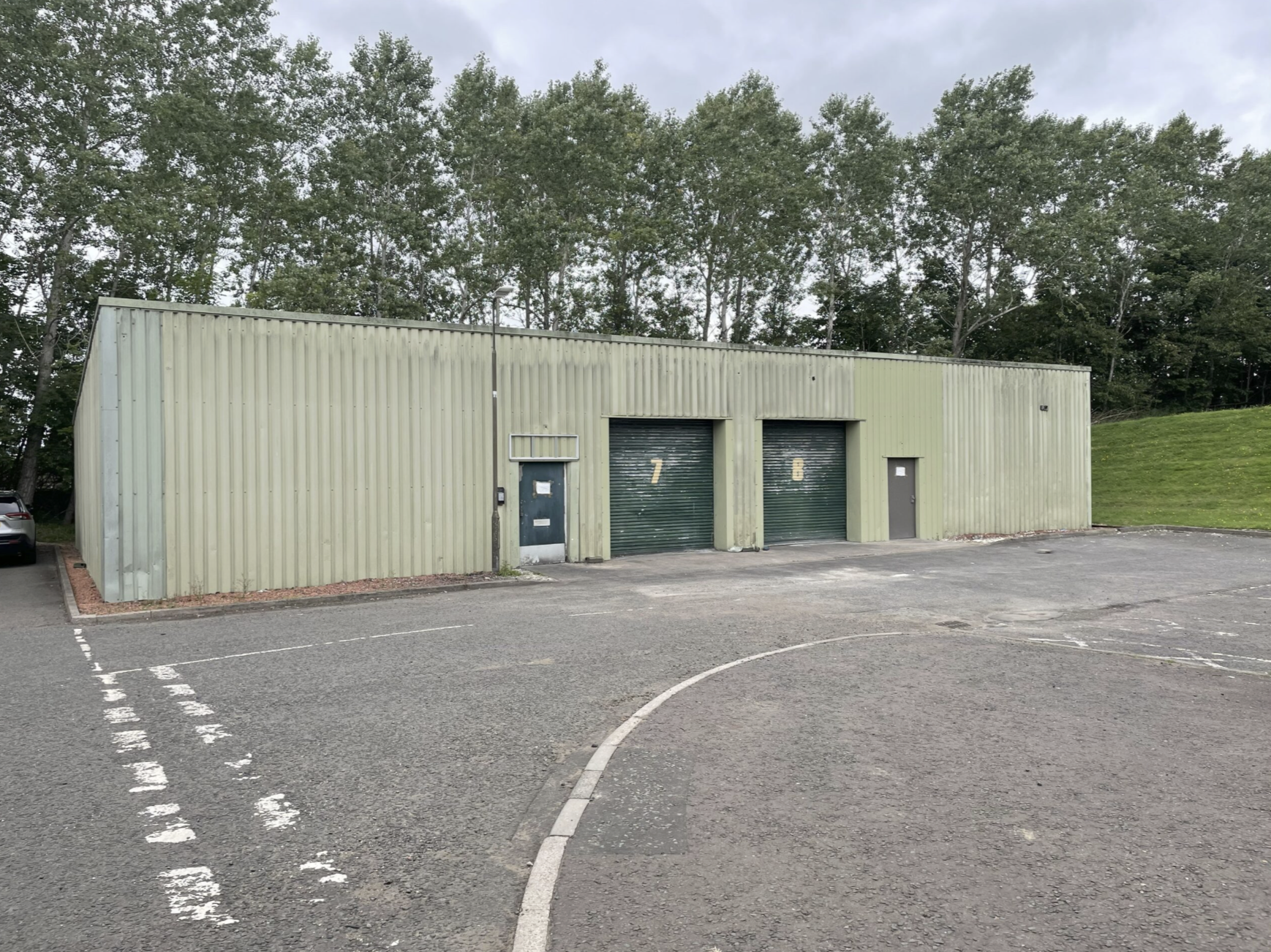 An industrial building with two green roll-up doors numbered 7 and 6, a small gray door, and a parking lot with marked spaces, some cars, and a grassy area with trees in the background.