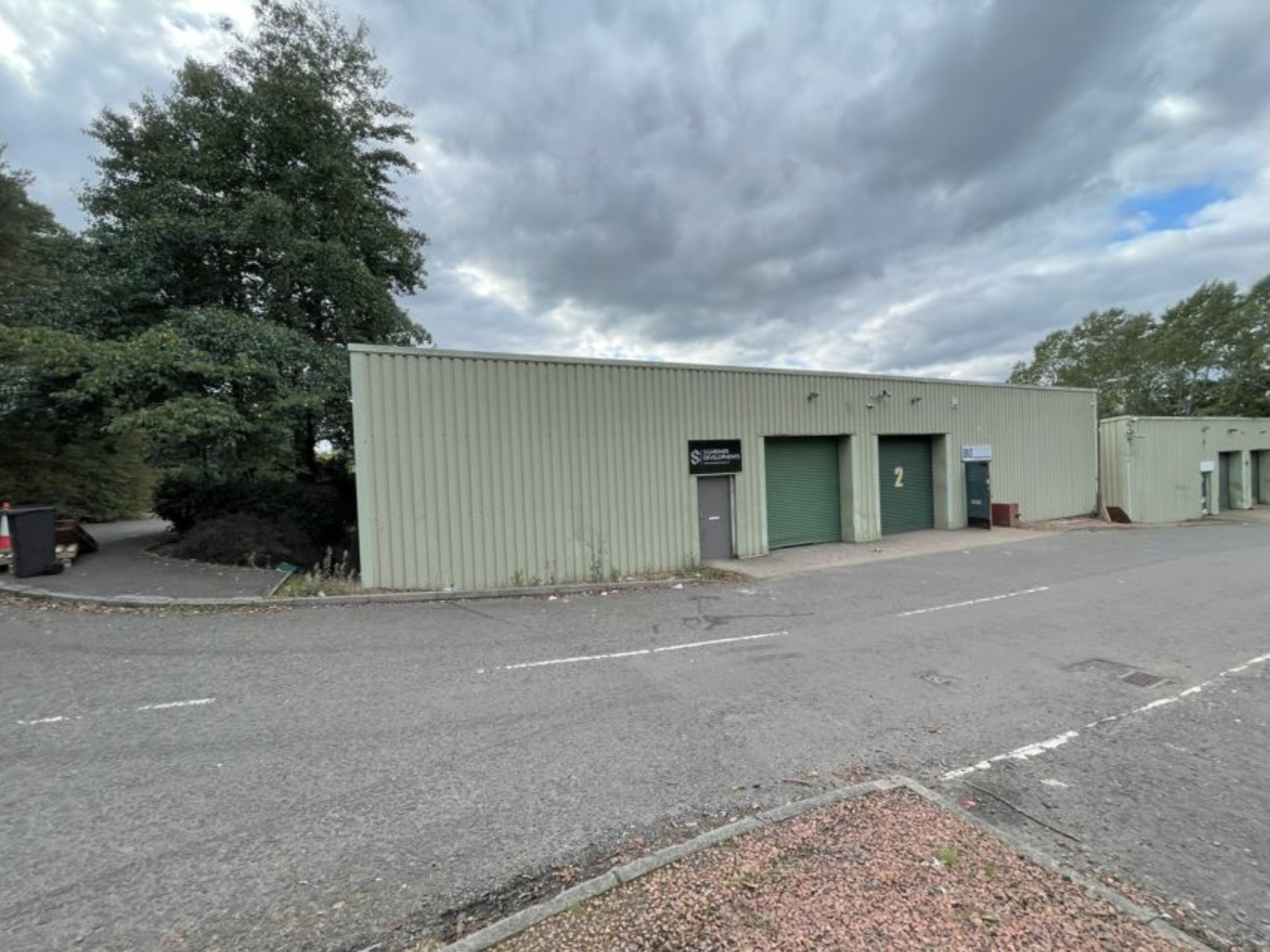 A beige industrial building with two green roll-up garage doors and a small door in between, situated in a parking lot with a cloudy sky overhead.