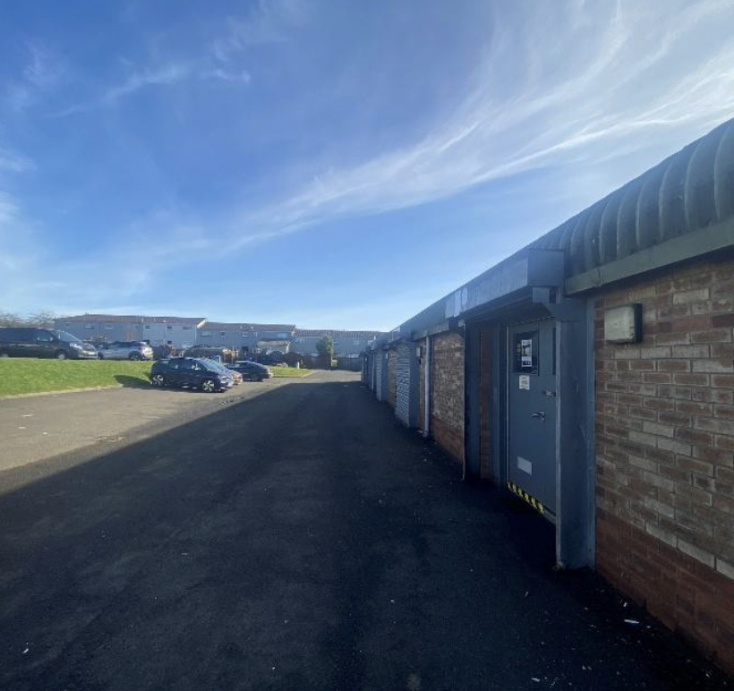 A parking lot with several parked cars, a building with brick and metal doors, and a blue sky with scattered clouds.