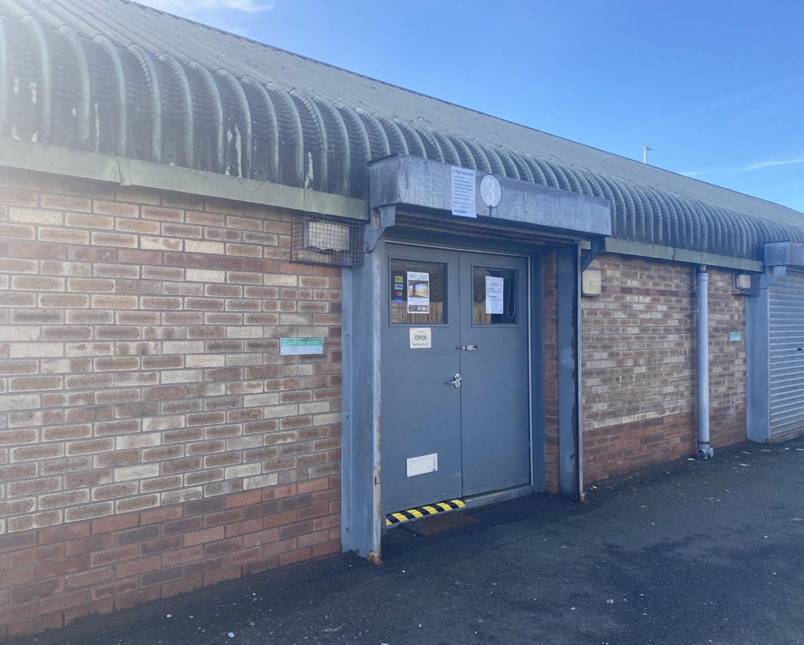Brick building with gray double doors, small windows, and a metal overhang. The ground is asphalt, and the sky is blue with few clouds.