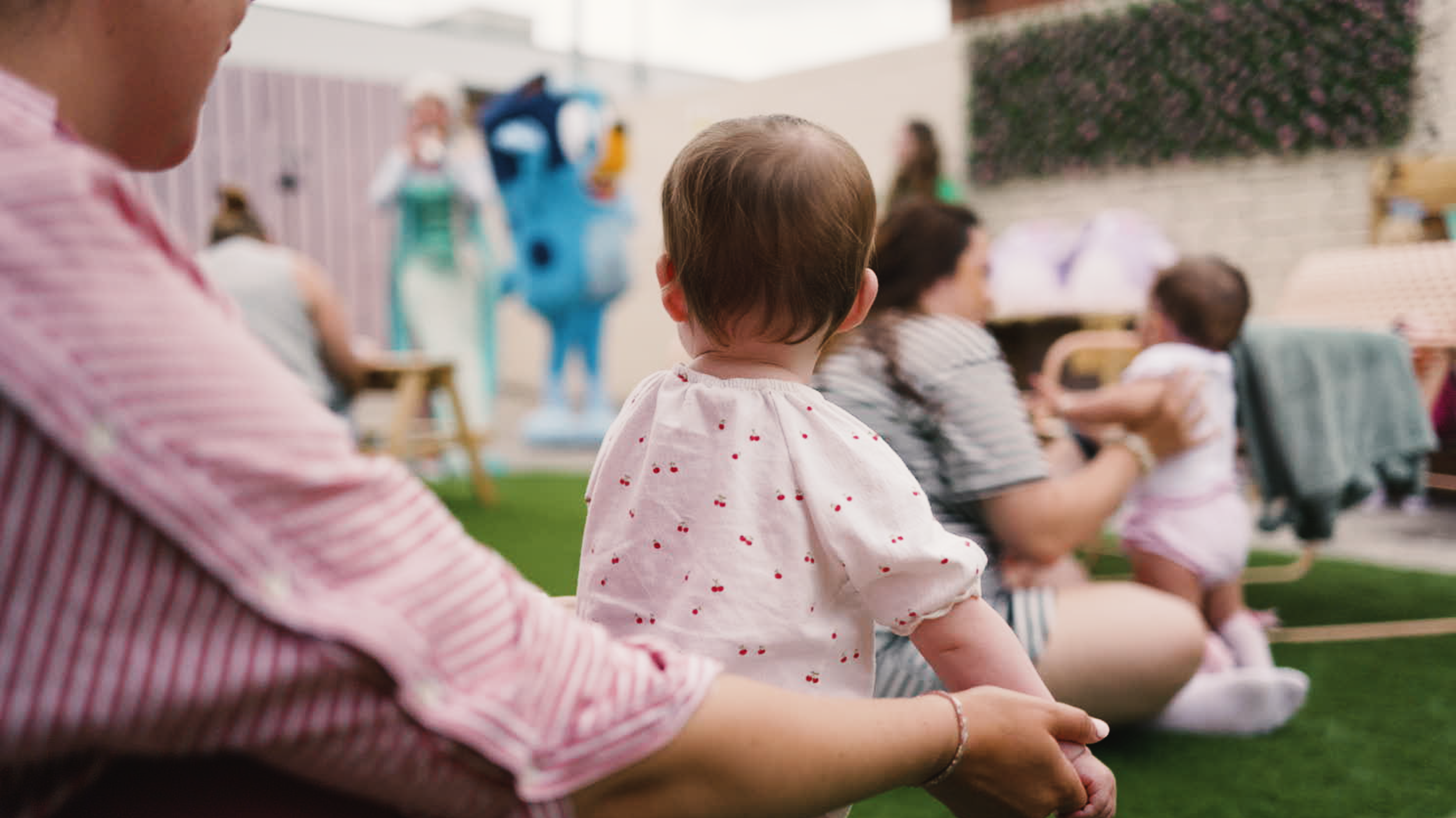 Children and adults seated outdoors, watching an event with a person dressed in a blue mascot costume in the background.