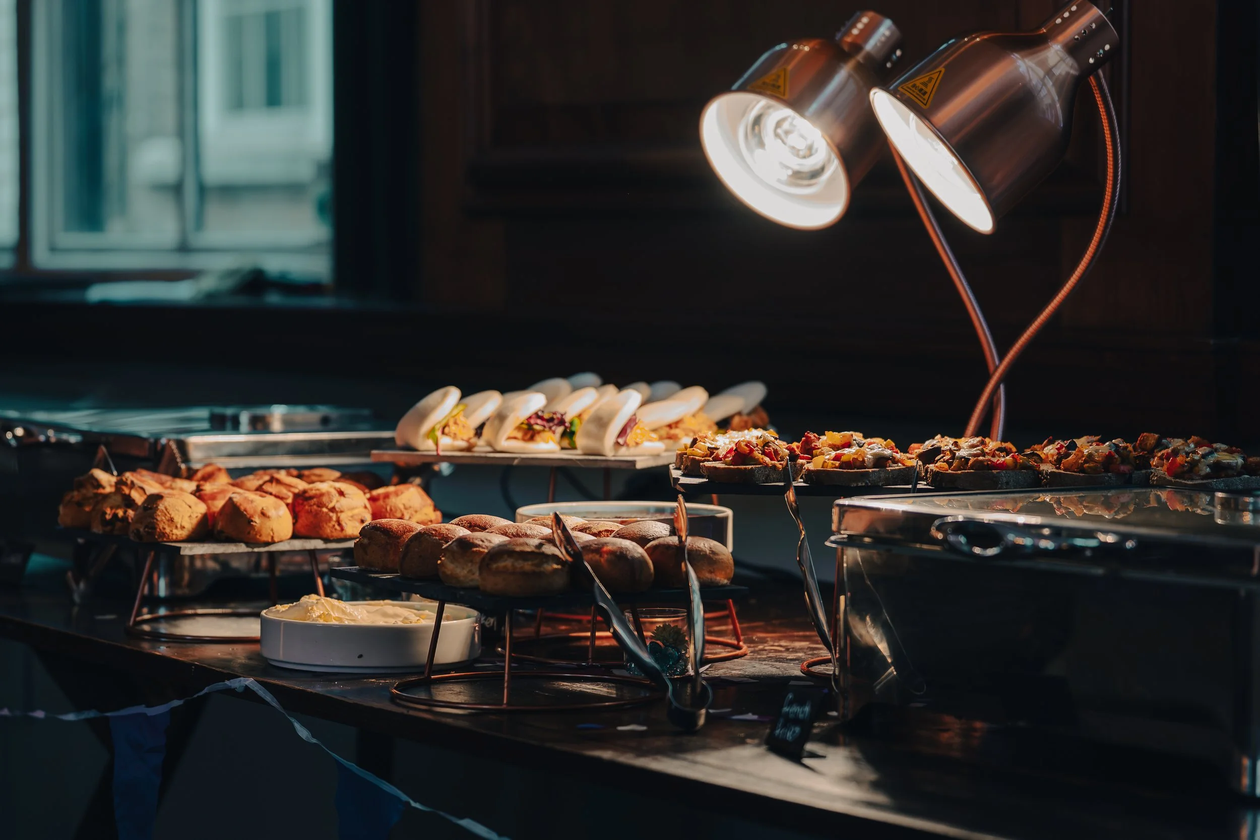 Buffet table with various food items, including sandwiches, pizzas, baked goods, and a bowl of dip, under warm lighting.