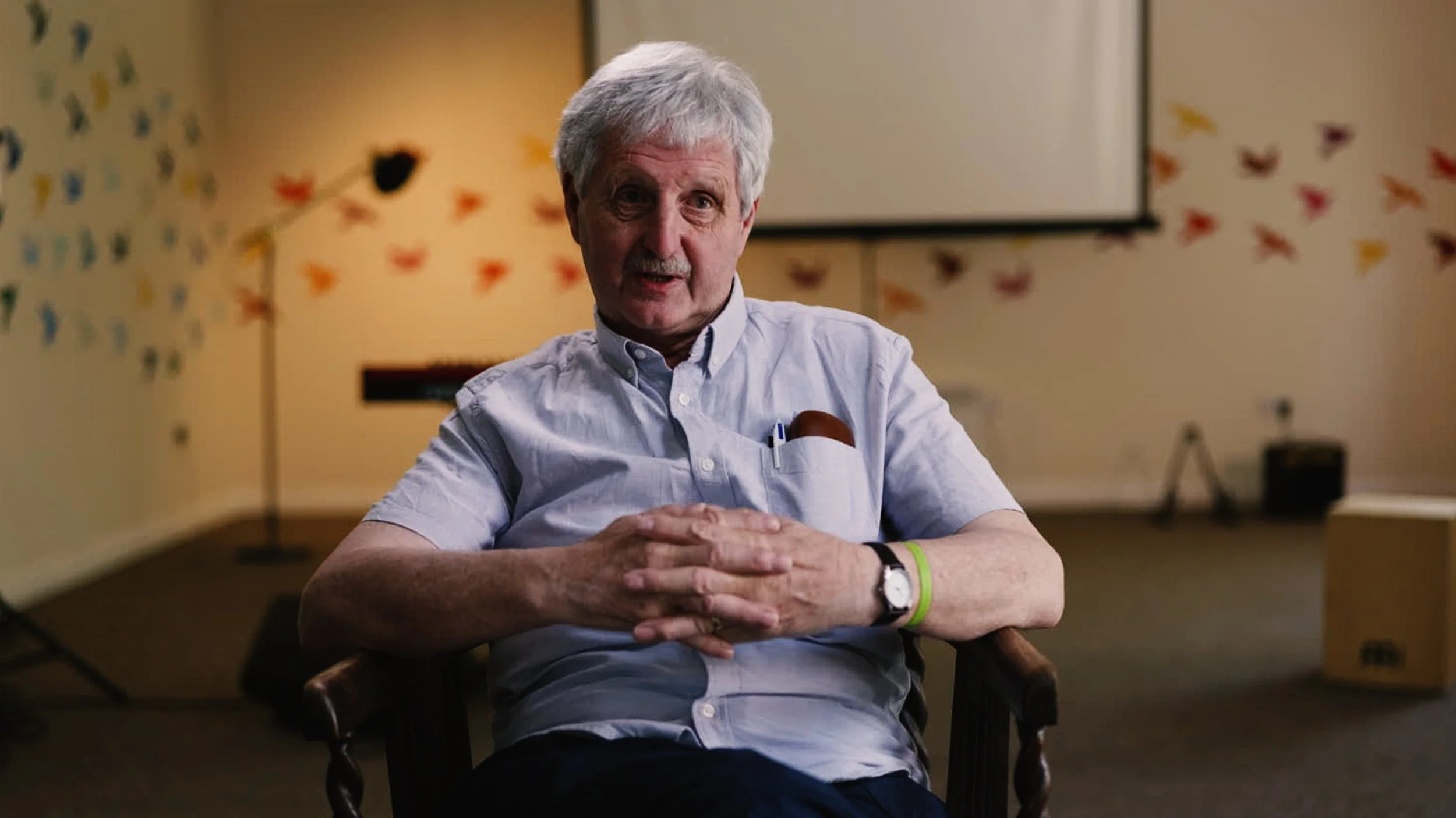 An elderly man with gray hair wearing a light blue short-sleeved shirt, sitting at a table with his hands clasped. He has a watch on his left wrist and a lime green wristband on his right wrist. The background features a beige wall with colorful butterfly decorations and a large white screen.
