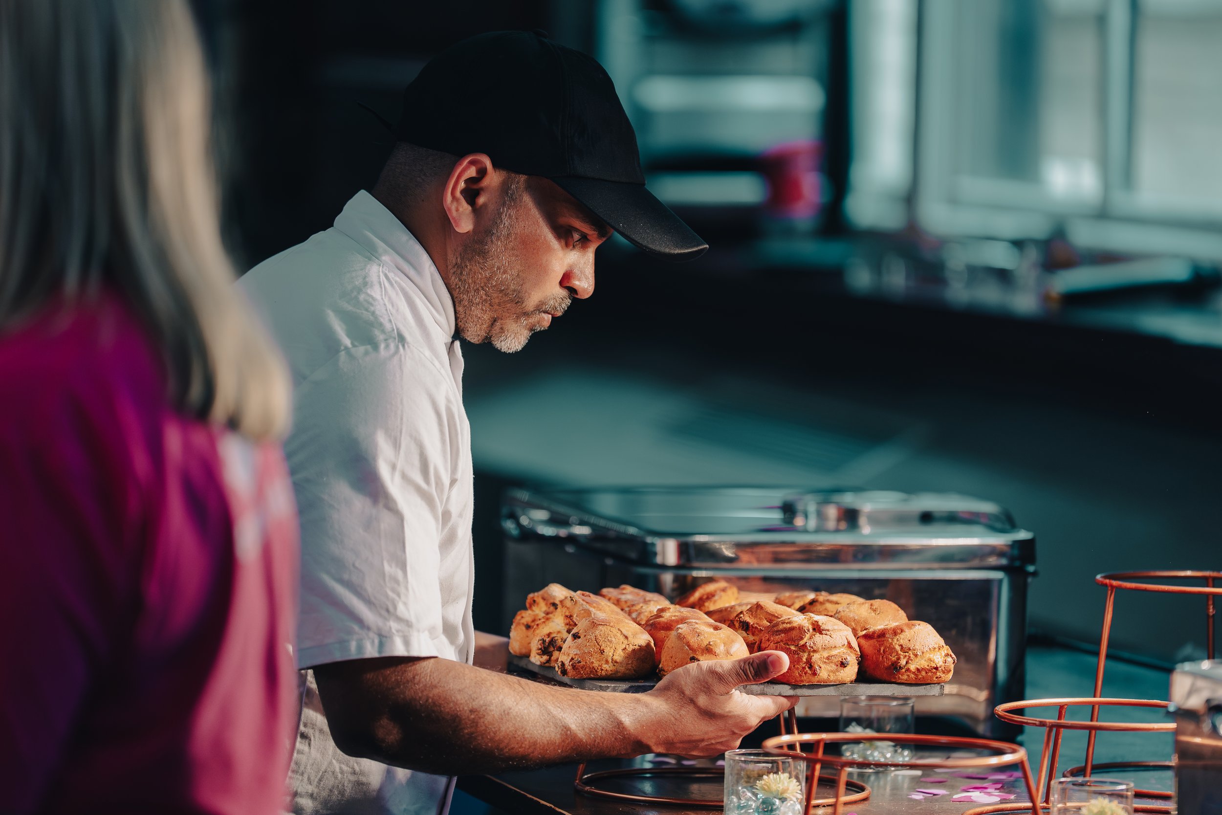 A man wearing a black cap and white chef's coat holding a tray of baked bread rolls in a bakery kitchen.