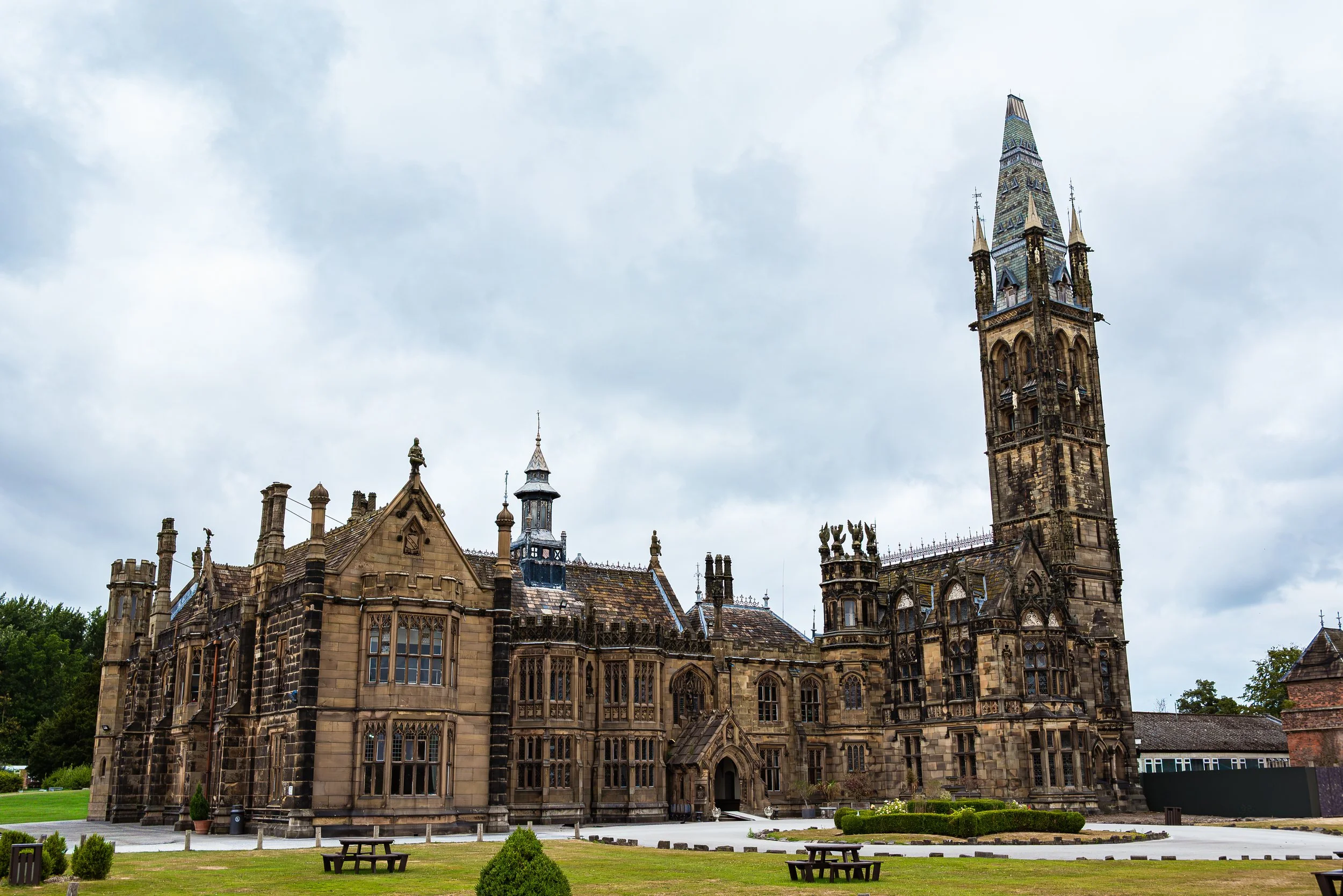 A historic Gothic-style building with intricate stonework and tall, pointed spires, located in a park with grass and benches, under a cloudy sky.