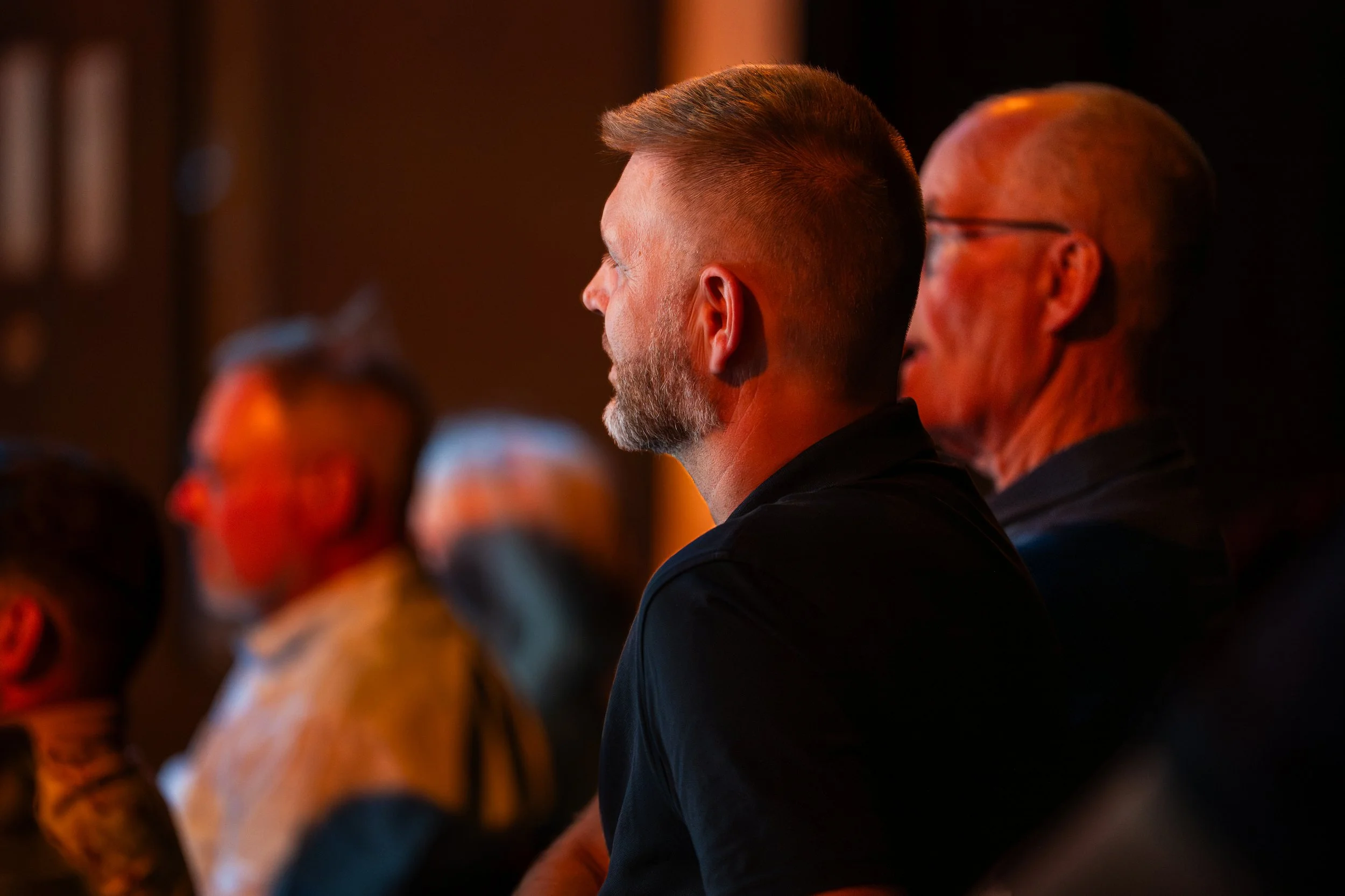Profile view of a man with short hair and a beard, wearing a dark shirt, sitting in a dimly lit room with blurred individuals around him.