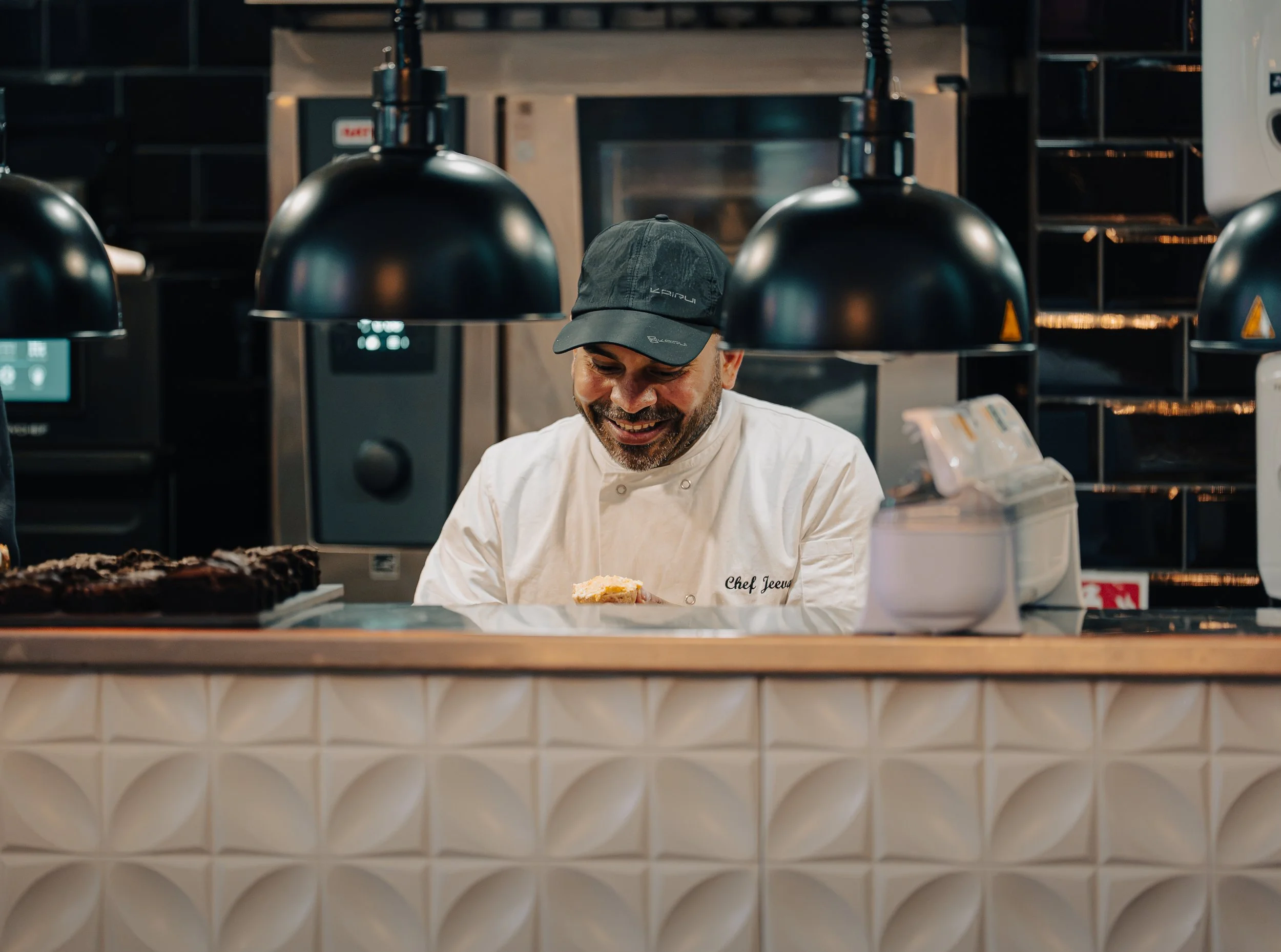 A chef with a grey cap smiling while working in a restaurant kitchen. There are black pendant lights overhead and baked goods or pastries on the counter in front.