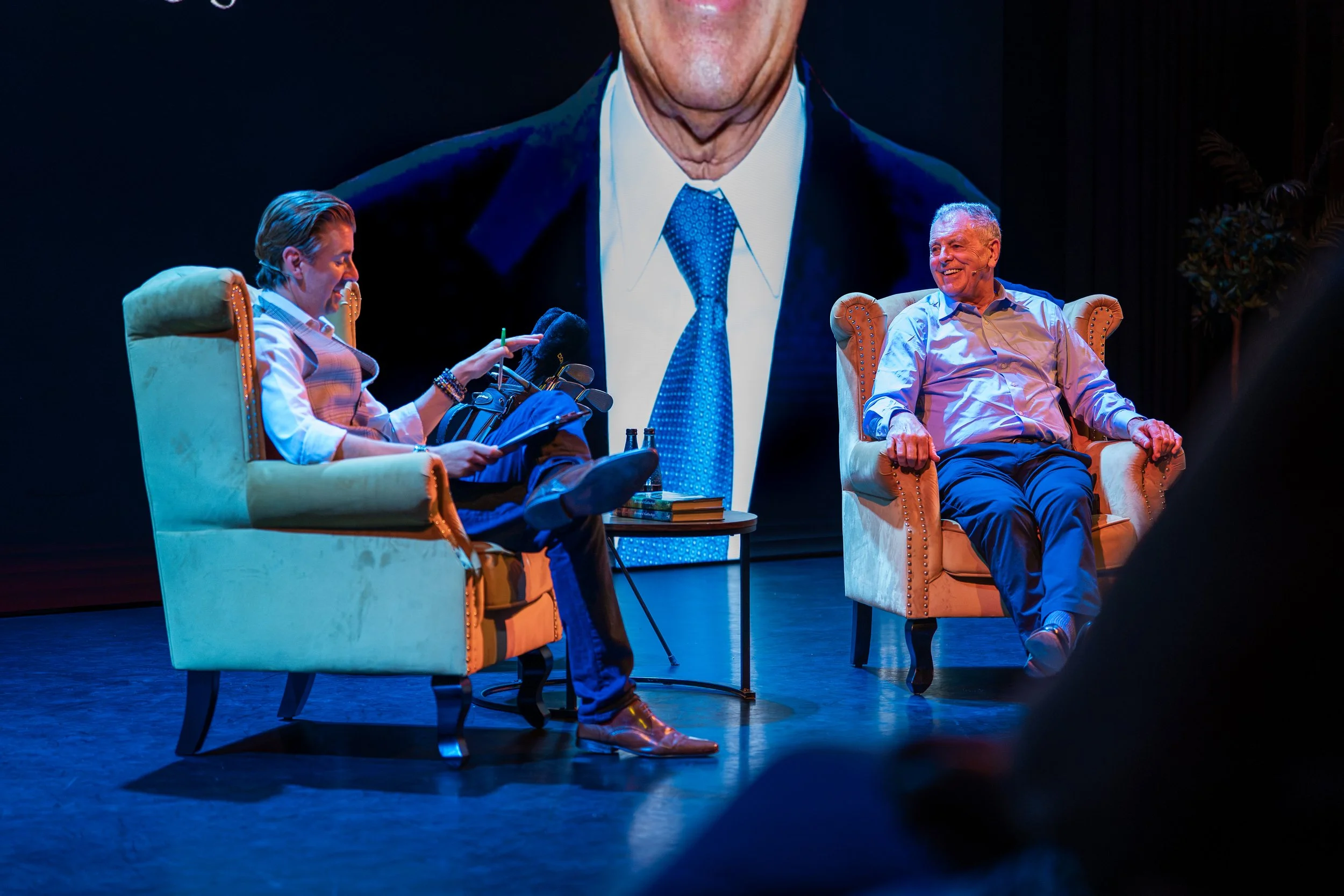 Two men are seated on stage chairs, engaging in conversation and laughing. A large digital screen behind them displays a close-up of one of the men's faces. The stage is dimly lit with a dark background.