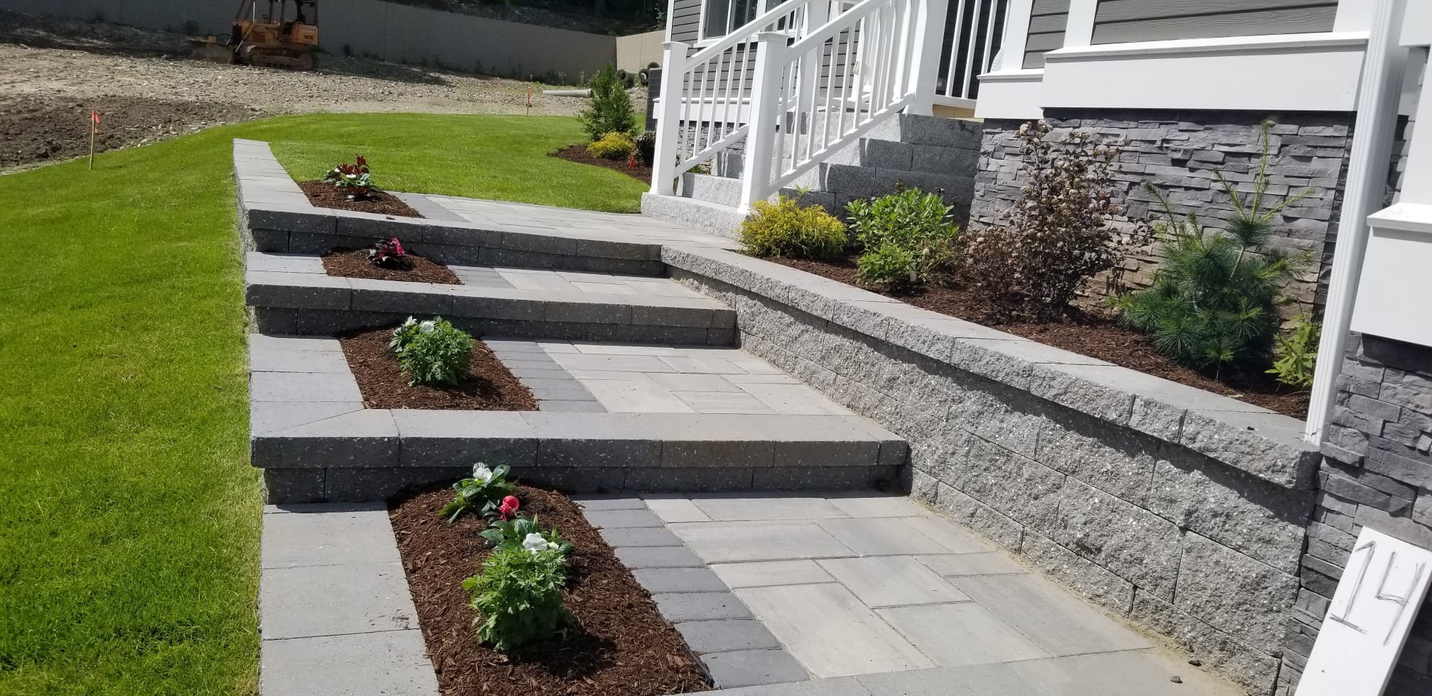 Freshly landscaped front yard with stone pathways and steps, green grass, and planted shrubs along a house wall.