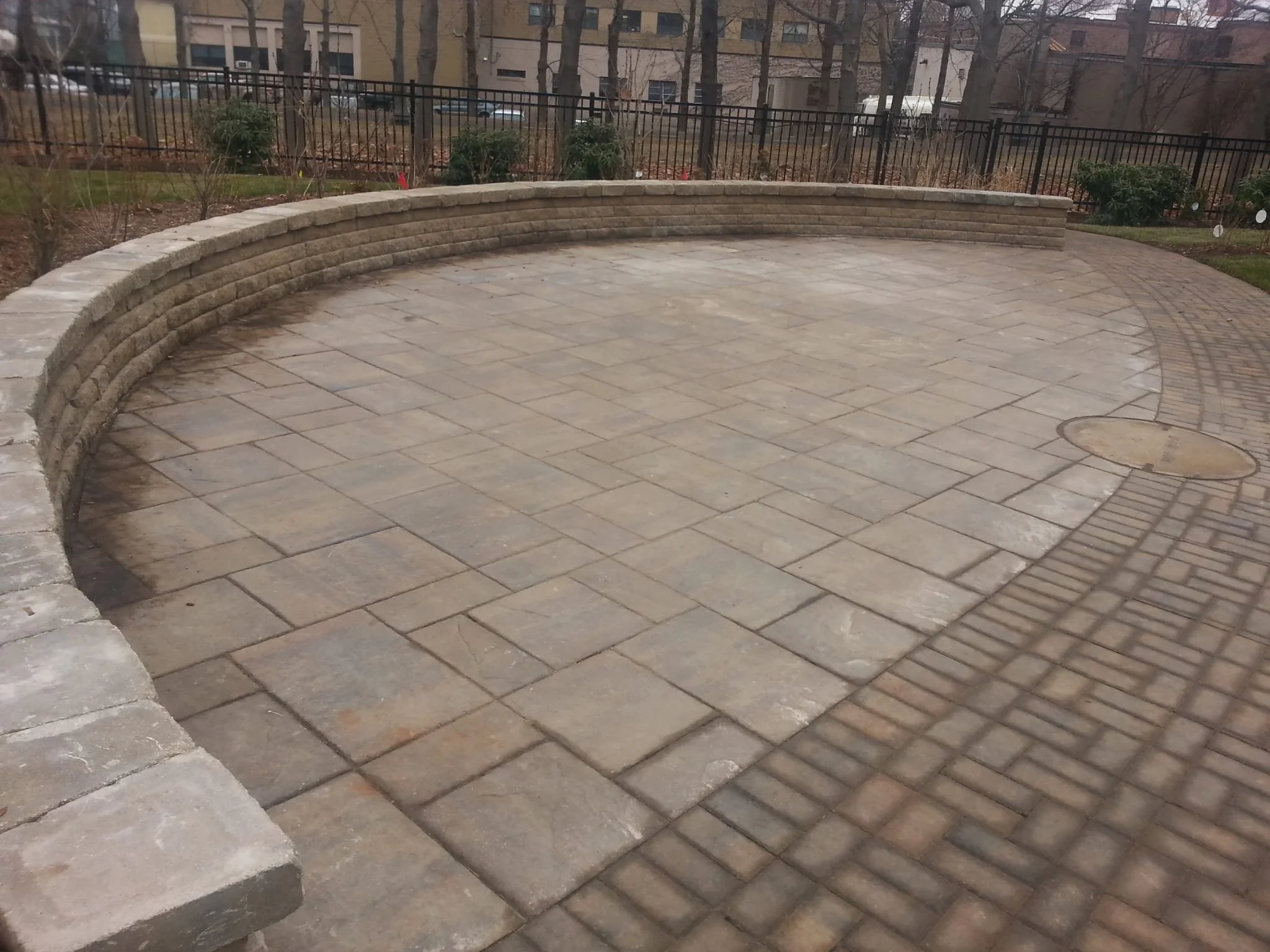 A paved outdoor patio area with a curved stone wall and a black metal fence, surrounded by shrubs and trees in a residential neighborhood.