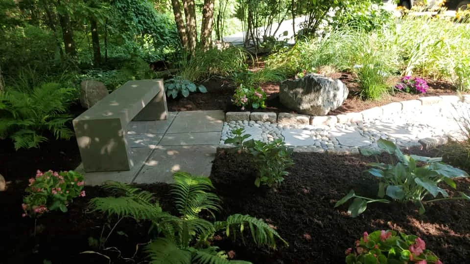 A garden with a concrete bench on a paved walkway, surrounded by lush plants, flowers, and rocks, with trees in the background and sunlight filtering through the foliage.
