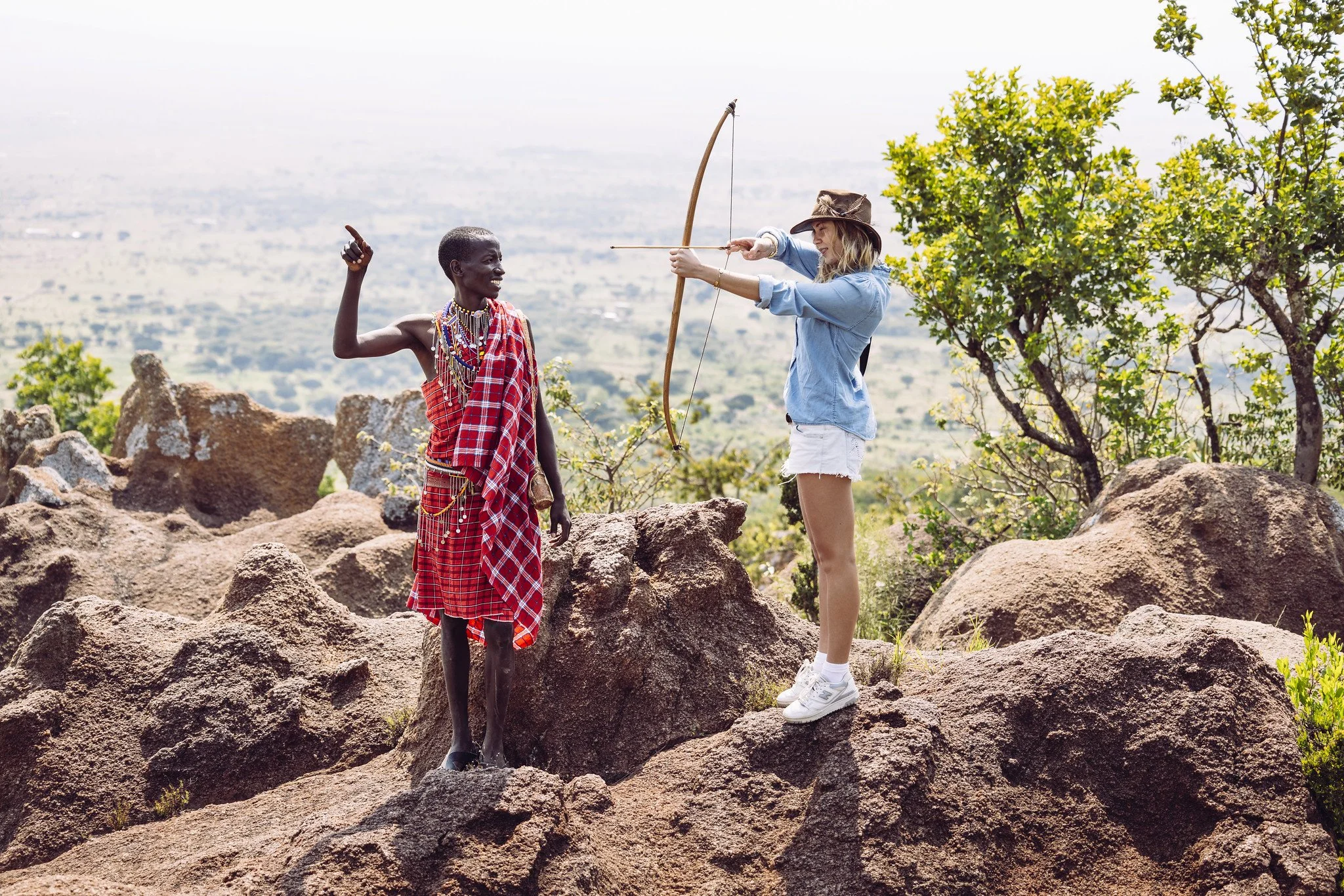A woman with blonde hair wearing a hat, light denim jacket, white shorts, and white sneakers is aiming a bow and arrow at a Maasai woman dressed in traditional red and plaid shuka with beaded jewelry. They are standing on rocky terrain with green tre