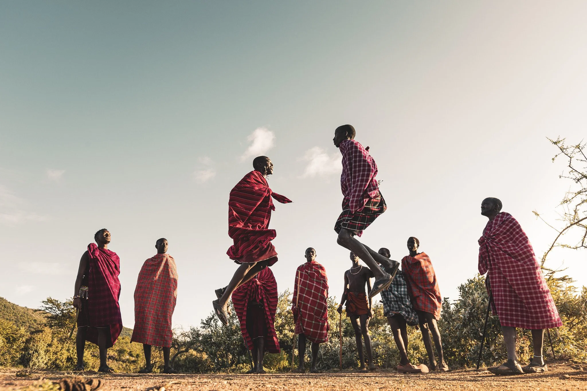 Maasai men and women in traditional red shuka cloth, some jumping and some standing, outdoors against a clear sky at sunset in a rural landscape. Maasai culture safari experience. Bucket list itinerary Kenya.